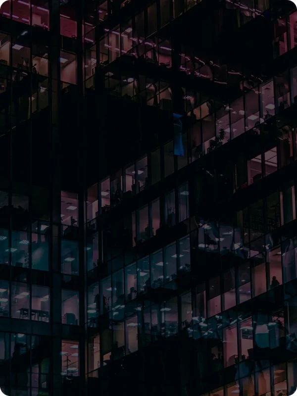 Nighttime view of a modern glass office building with illuminated interior offices and workplaces visible through the windows.