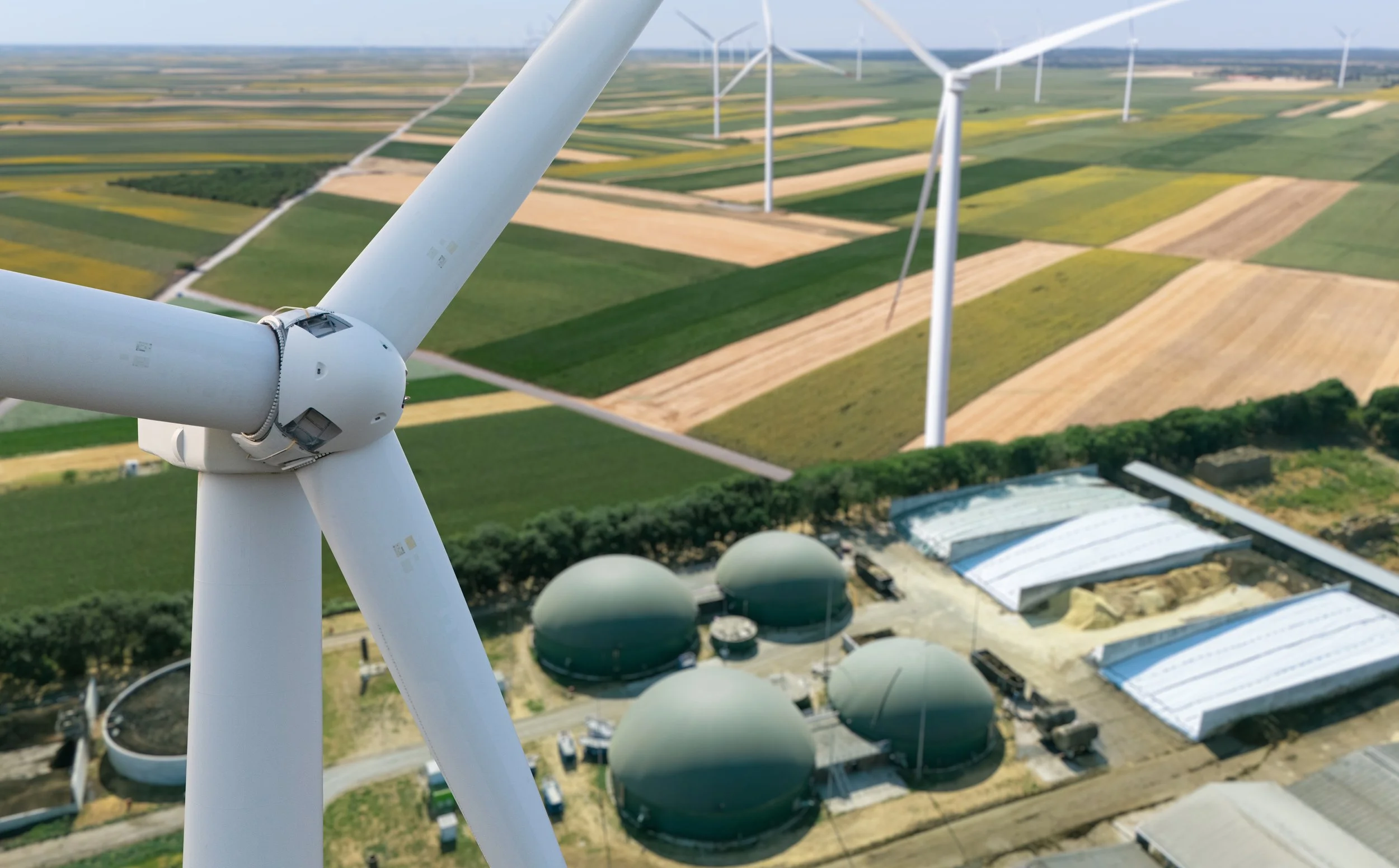 A close-up view of a wind turbine with several other wind turbines in the background in a rural landscape with patchwork fields and some industrial structures.
