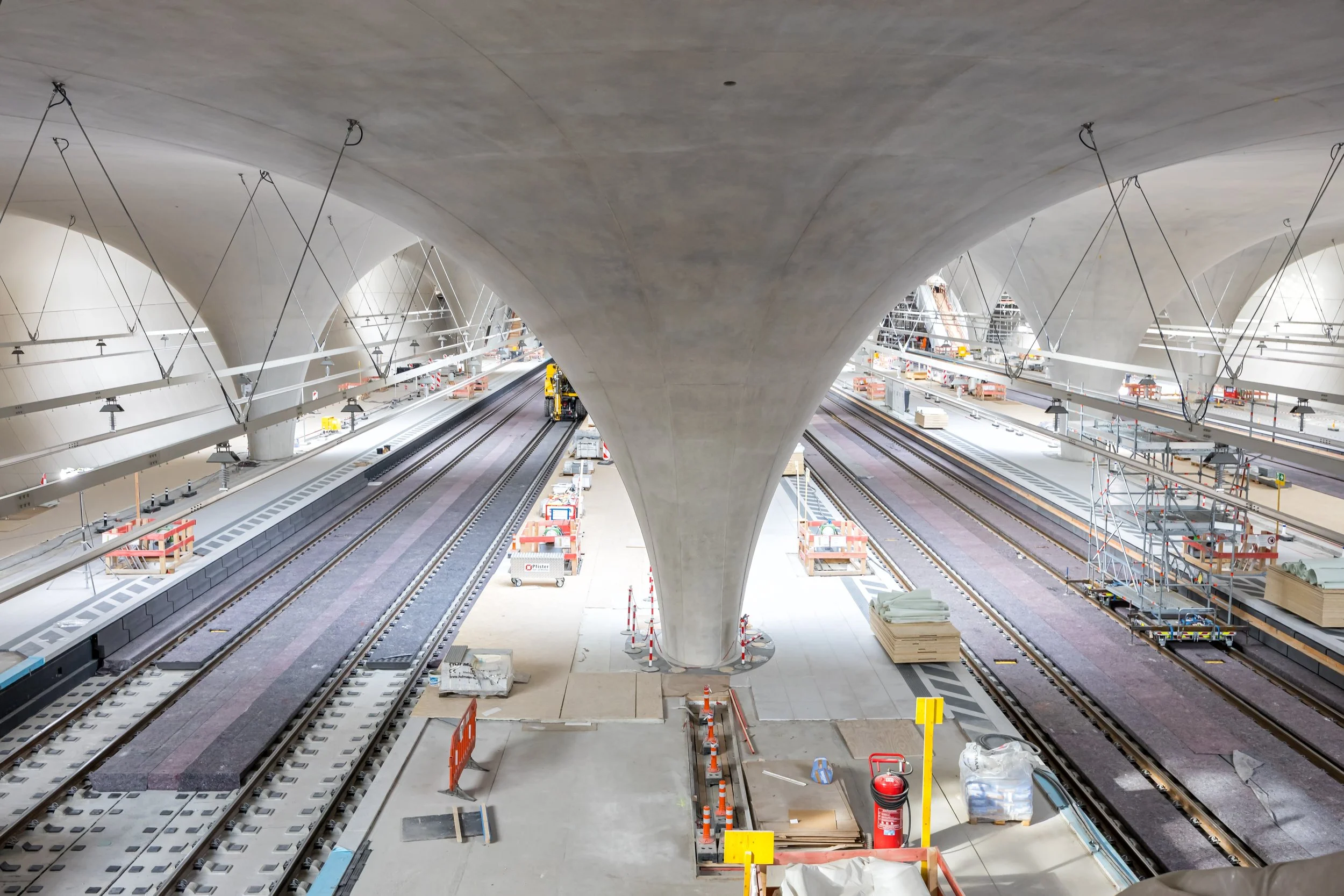 Underground train station construction site with railroad tracks, large concrete columns, and construction materials.
