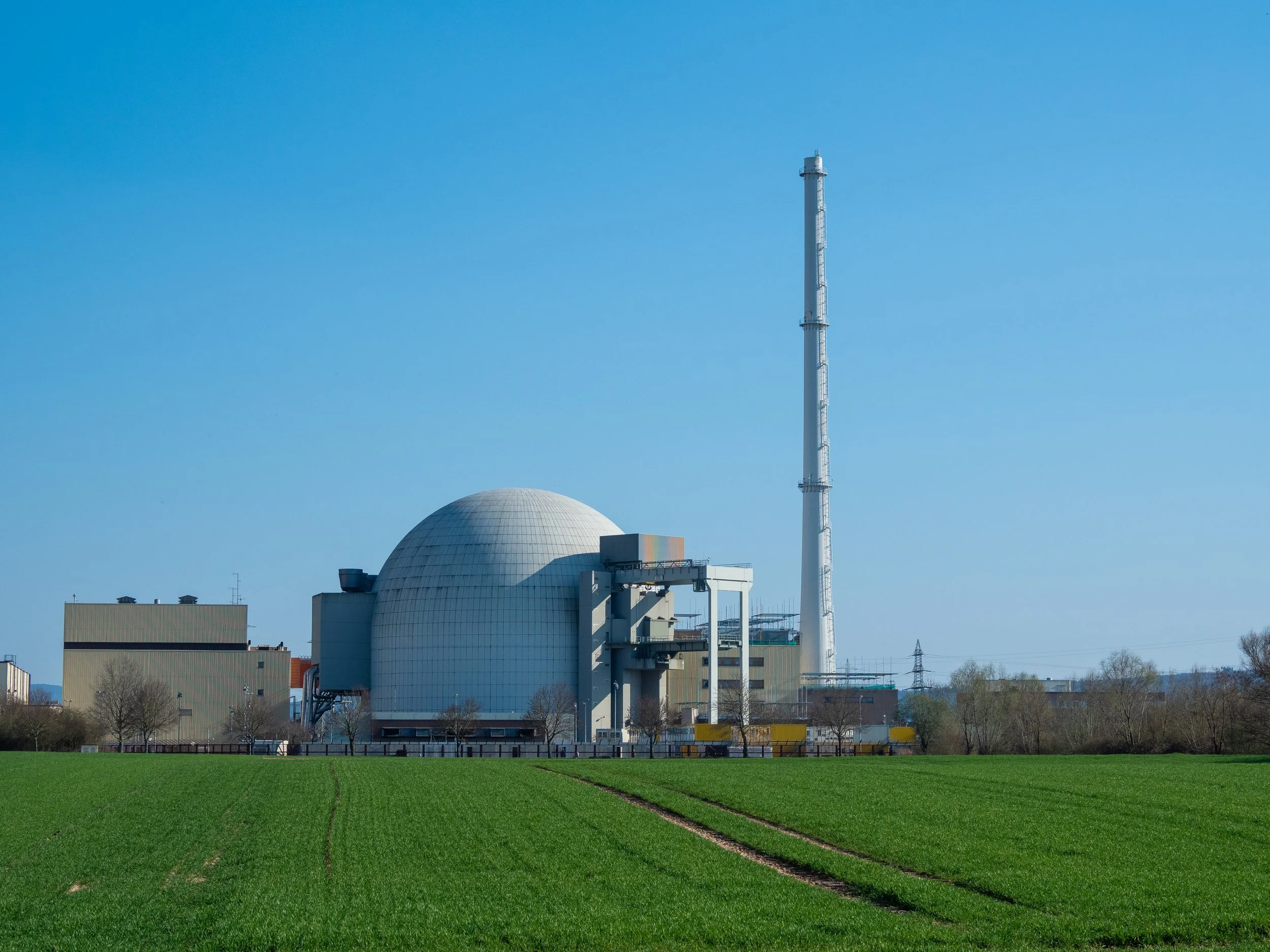 A large industrial building with a white domed cooling tower and a tall grey telecommunications tower against a blue sky, with a green field and a few trees in the foreground.
