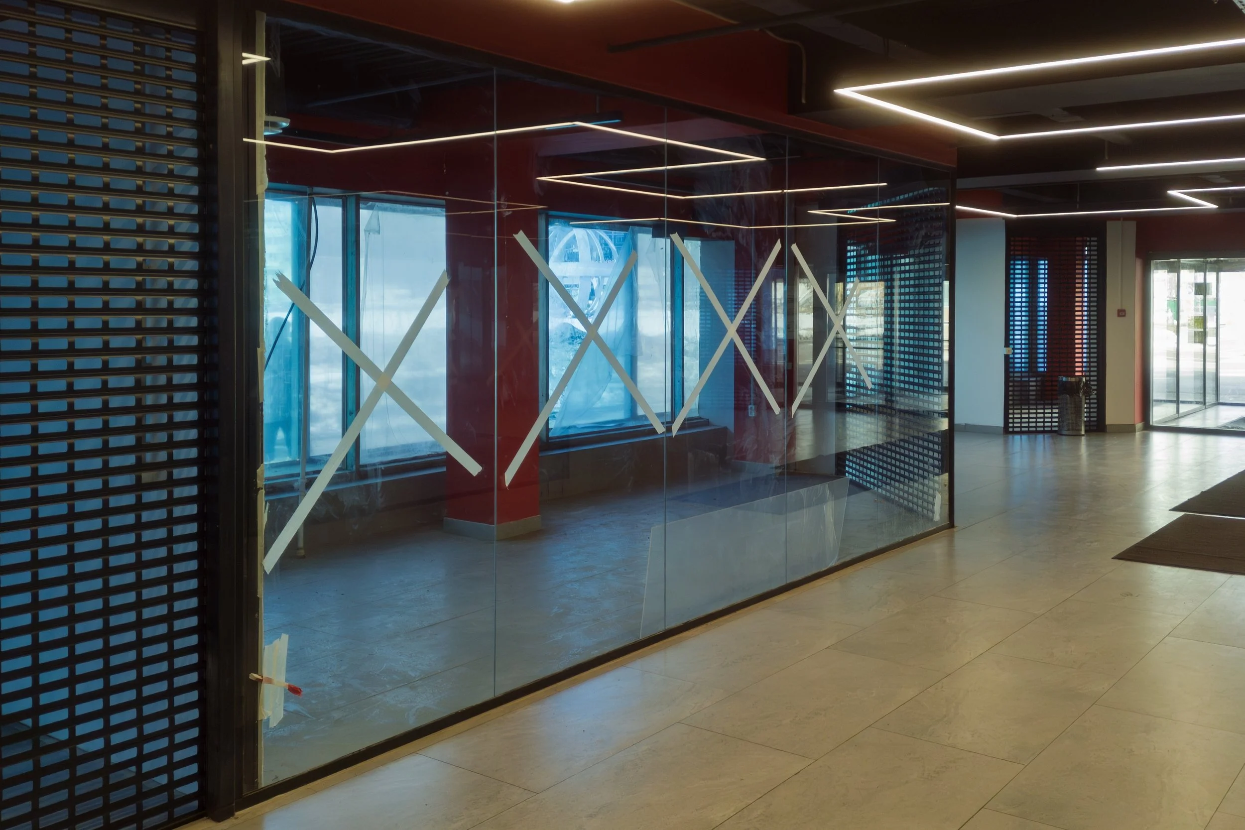 Modern interior space with glass walls, horizontal light fixtures on the ceiling, and a mix of natural and artificial lighting. Features include a glass door, black grid panel, and polished tiled floor.