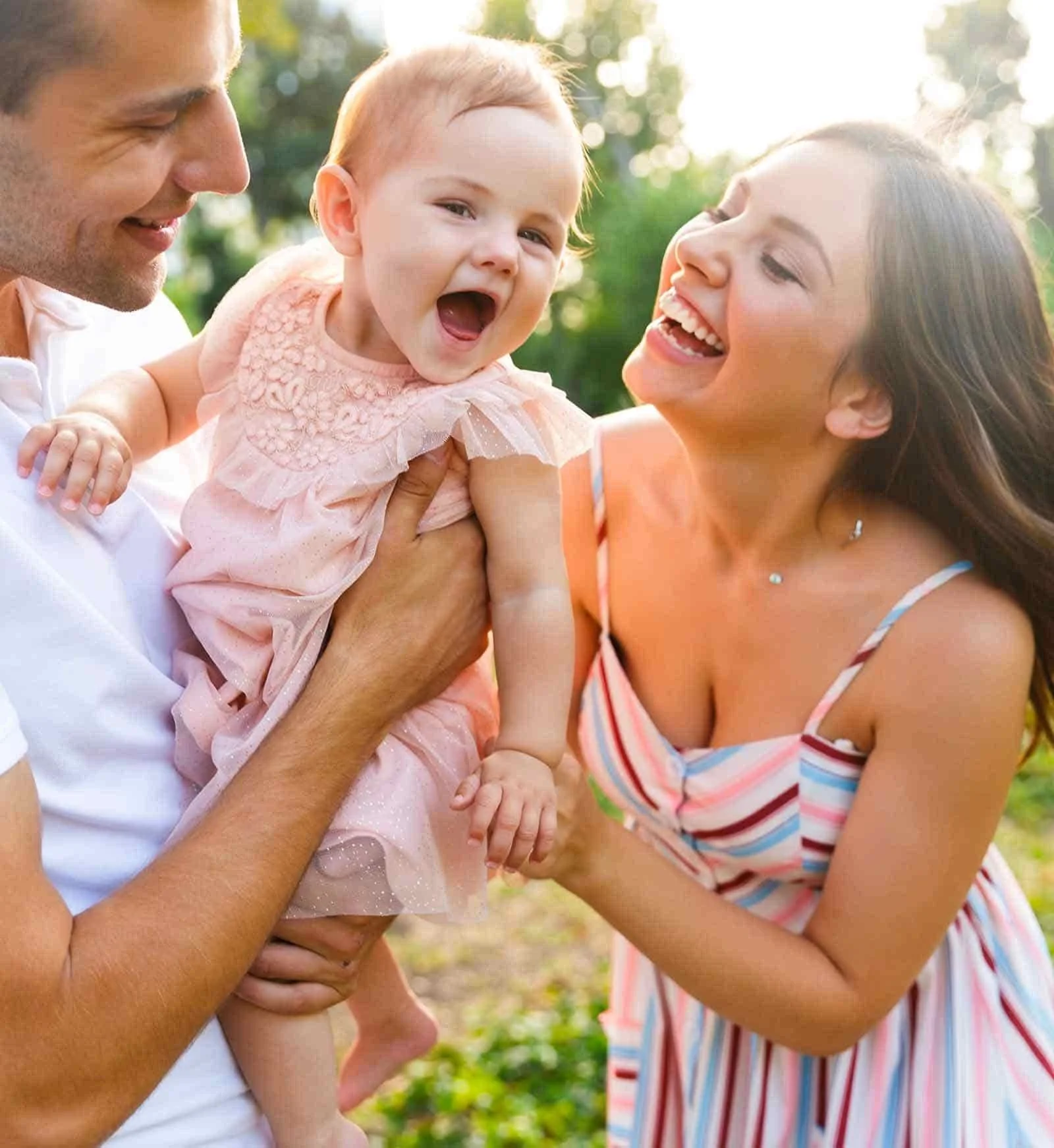 Une famille souriante en plein air, composée d'un homme, d'une femme et d'un bébé. L'homme tient le bébé, qui rit, tandis que la femme sourit vers eux. La lumière du soleil filtre à travers les arbres en arrière-plan.