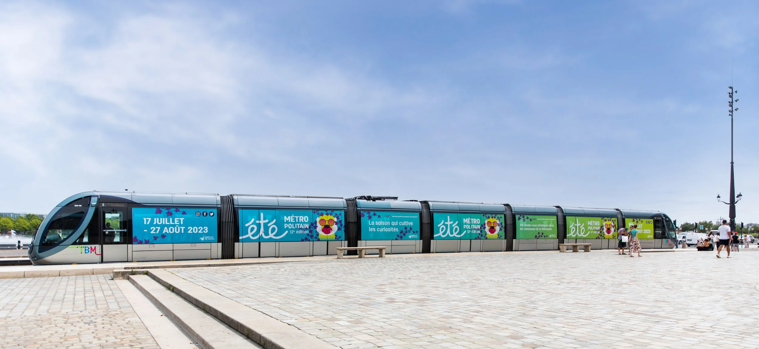 Un tramway moderne stationne en plein air, décoré avec des affiches promotionnelles pour l'été 2023 à Paris, avec plusieurs personnes qui attendent ou se déplacent à proximité.