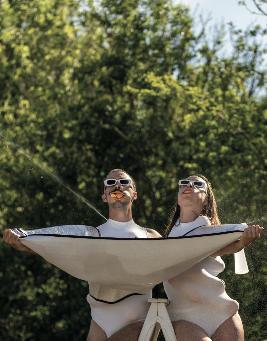 Deux personnes portant des lunettes de soleil blanches, avec des morceaux d'orange dans la bouche, tiennent une voile blanche dans un parc avec des arbres verts en arrière-plan.