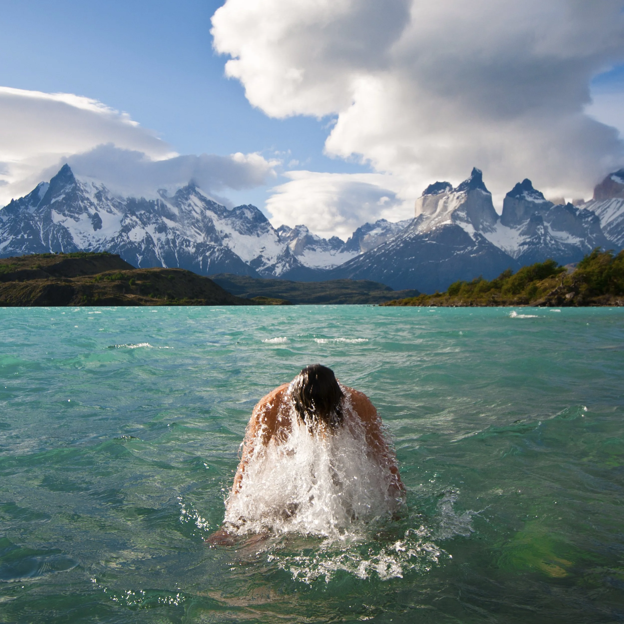 Une personne nage dans un lac turquoise avec des montagnes enneigées en arrière-plan sous un ciel partiellement nuageux — Gaspard Noël