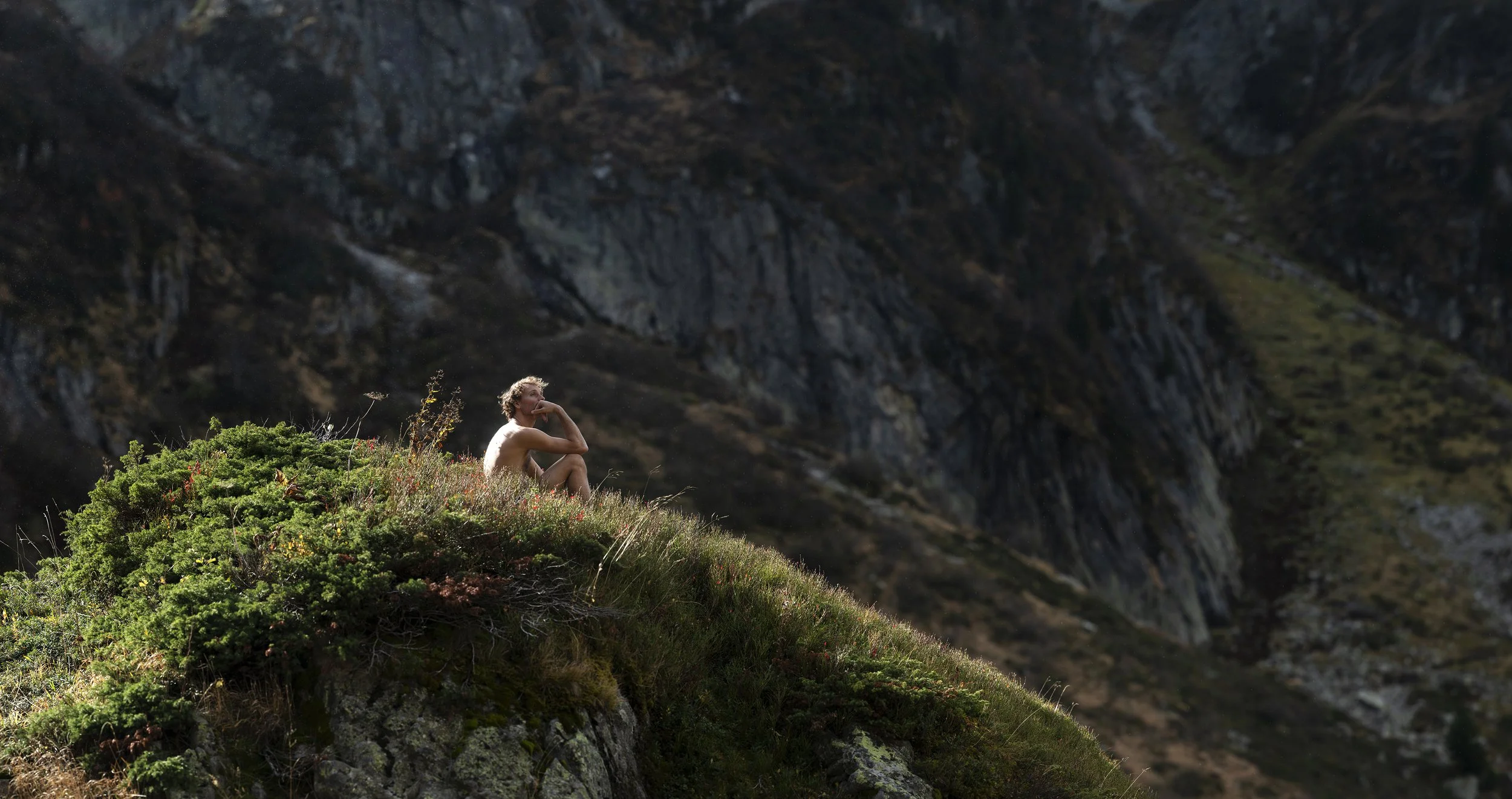 Une personne assise sur une colline recouverte de végétation, contemplant la nature avec des montagnes en arrière-plan. Thérapie.