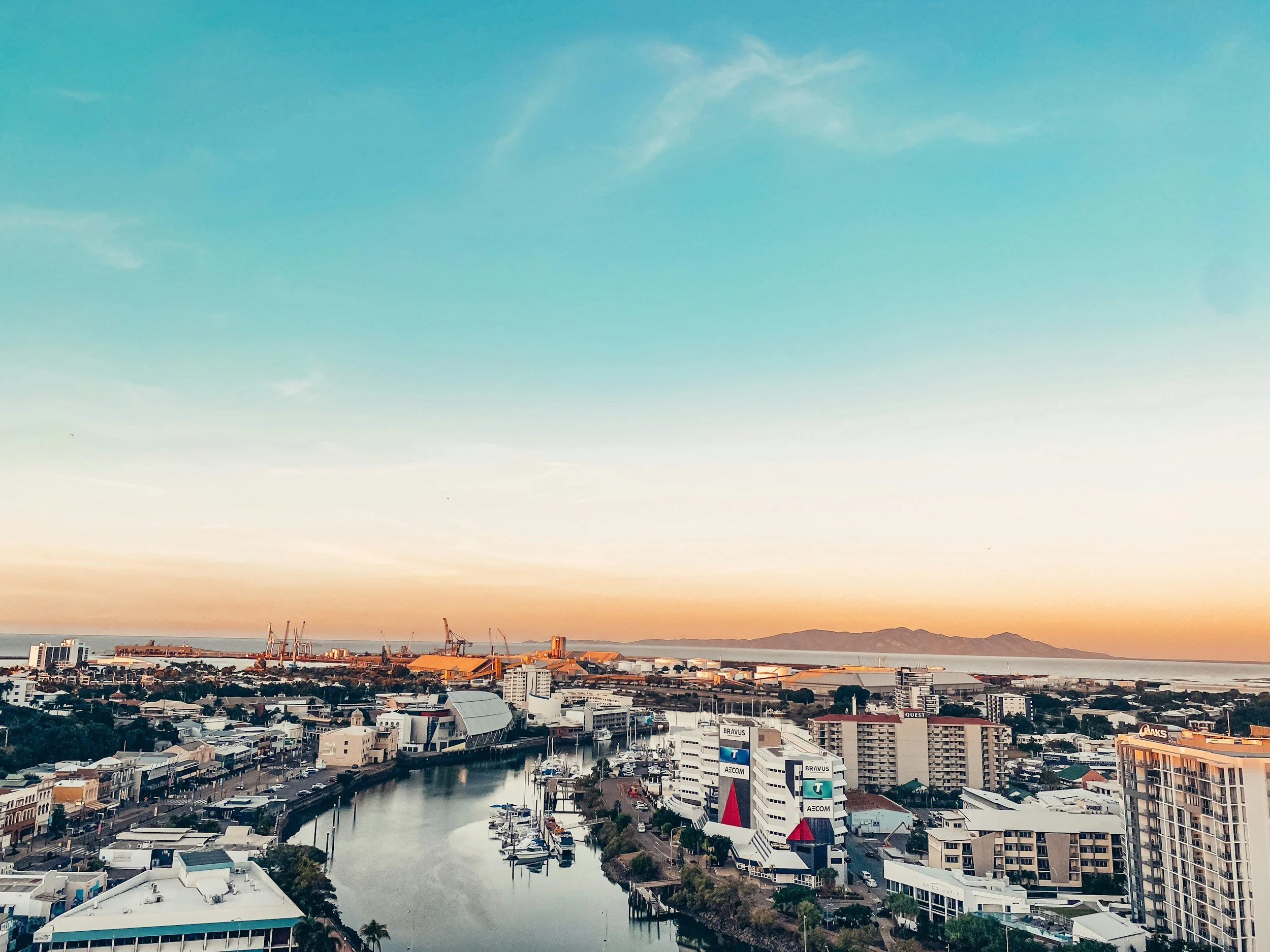 A view of the Townsville cityscape featuring buildings along a waterway with cranes in the background during sunset, under a blue sky with light clouds.