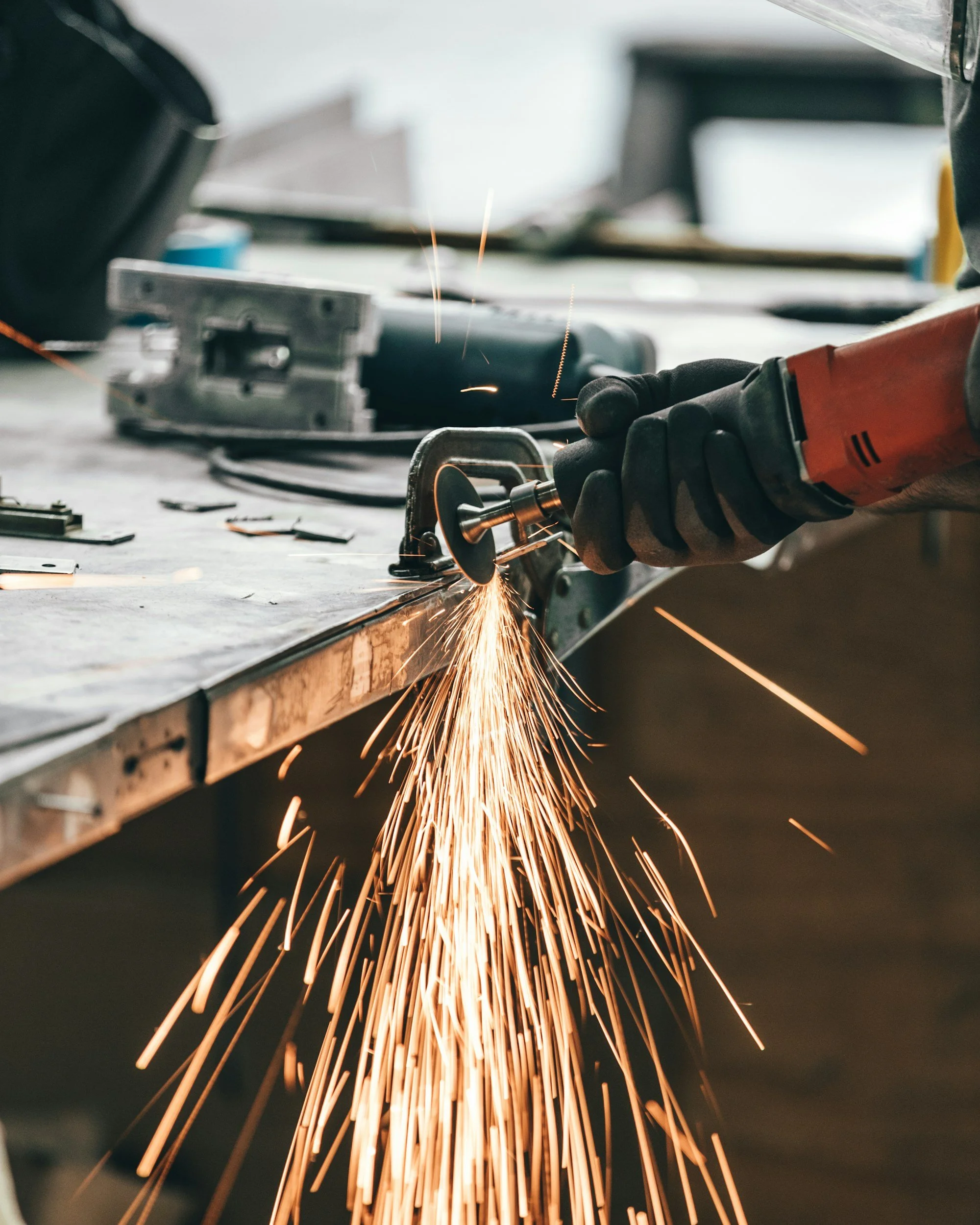 A person using an angle grinder to cut or grind metal, with sparks flying from the contact point, on a workbench in a workshop or industrial setting.