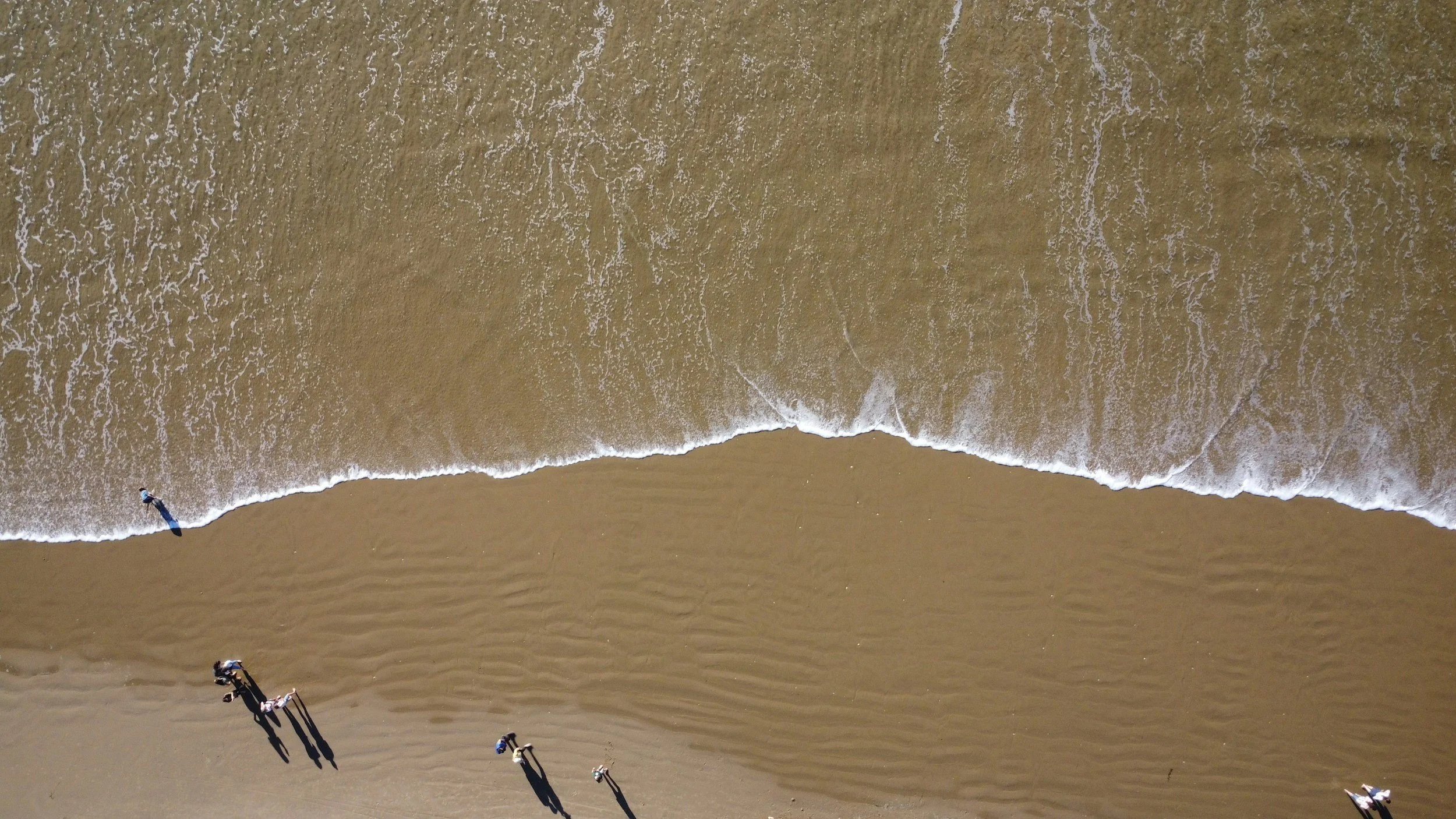Bird's eye view of a sandy beach with small waves and a few people walking along the shore.