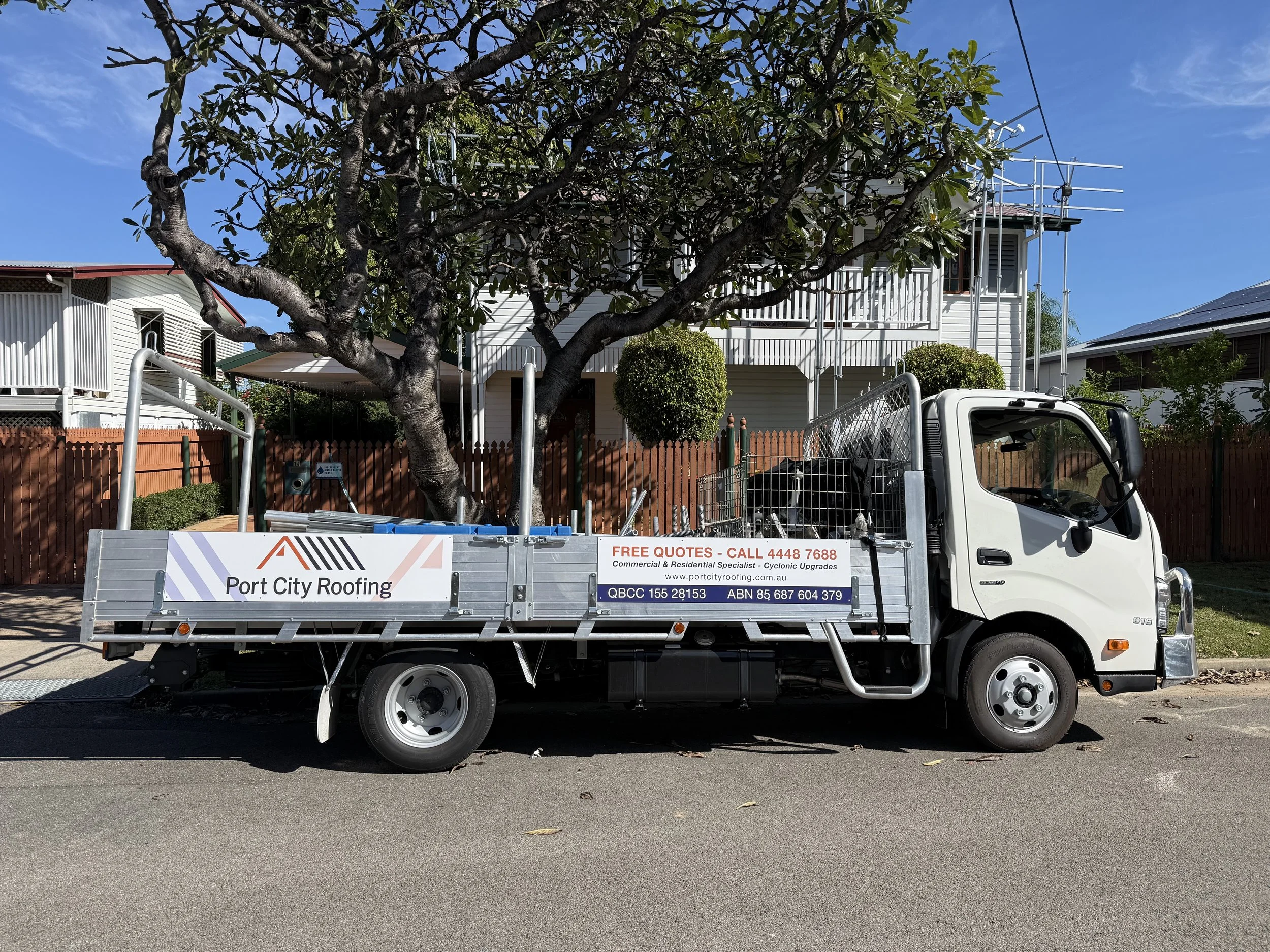 Port City Roofing's white Hino truck with a scaffolding setup on its bed parked in front of a residential house with white siding, trees, bushes, and edge rail errected.