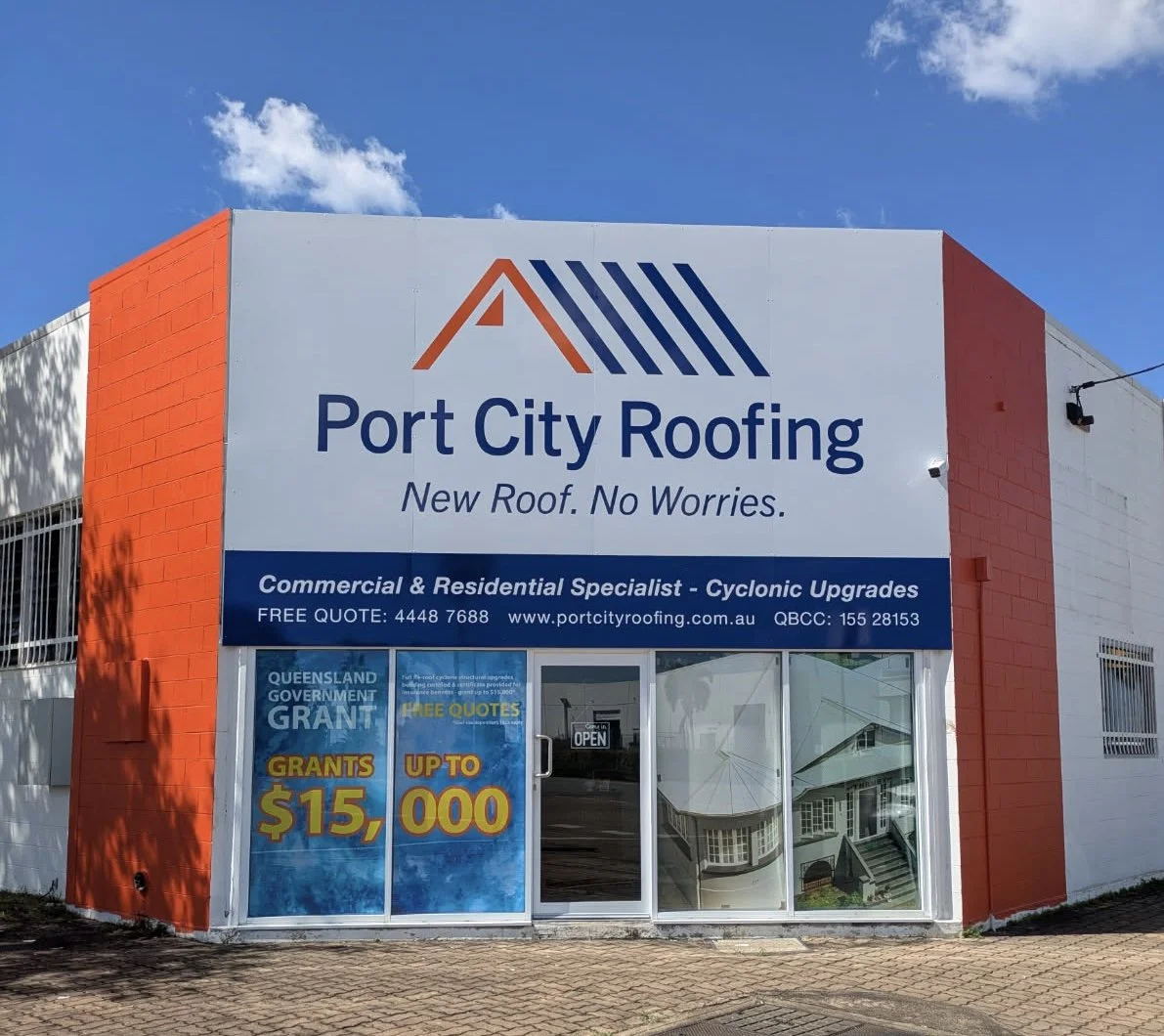 Exterio storefront of Port City Roofing with a logo, business name, tagline 'New Roof. No Worries.', and promotional posters indicating grants up to $15,000, with a storefront featuring an 'Open' sign and a large window reflecting the street.