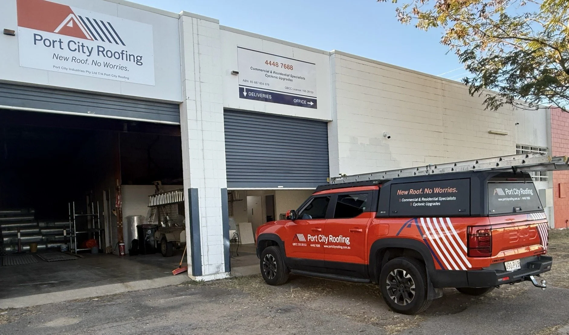View of Port City Roofing with a red service truck parked in front. The building has signs indicating roofing services and directions for deliveries and the office. The truck displays the company's logo, contact information, and services offered, including new roof installation and cyclonic upgrades.