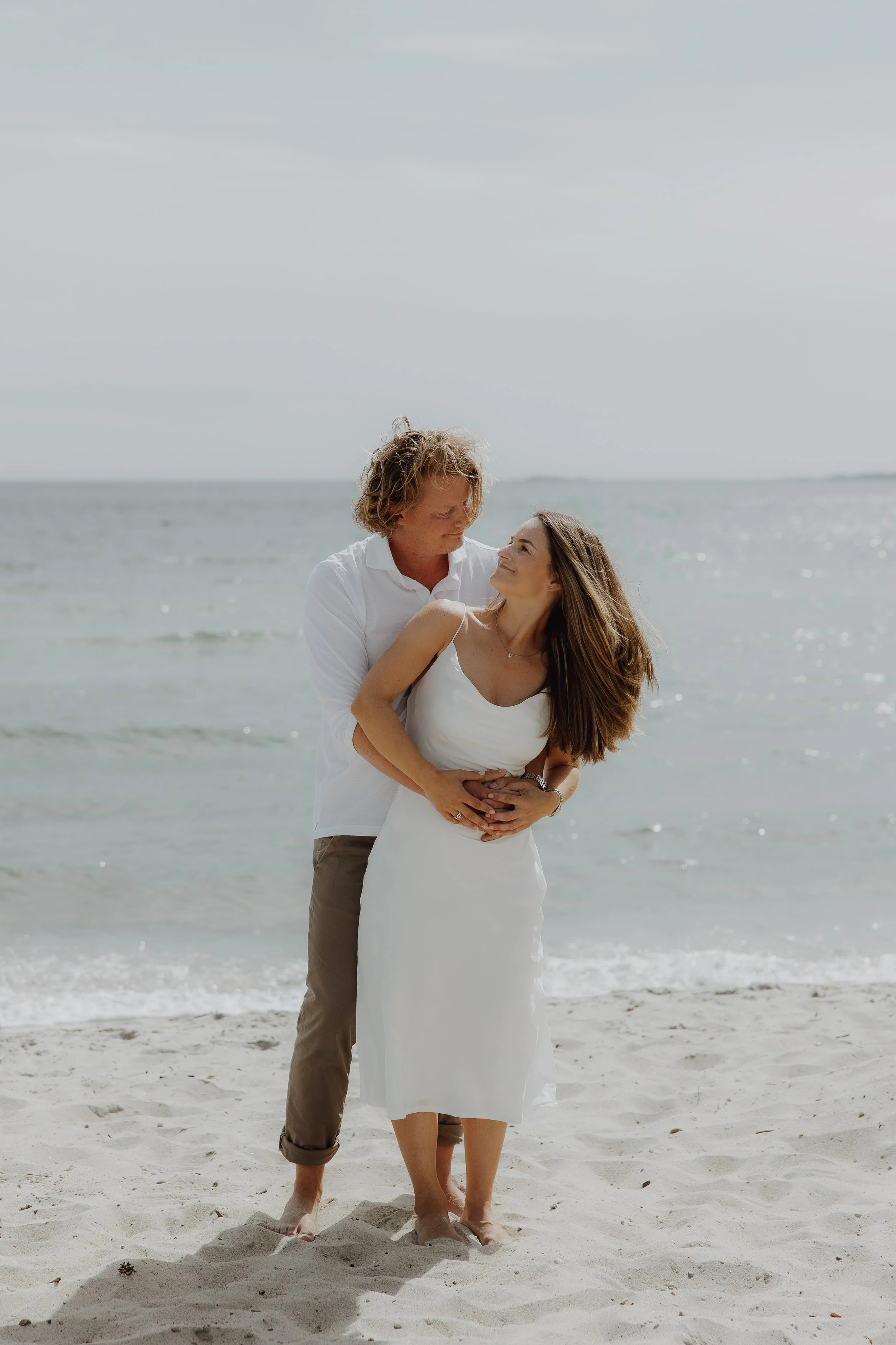 Et ungt par og ei jente står nær stranden, holder hverandre og ser på hverandre, med havet i bakgrunnen.