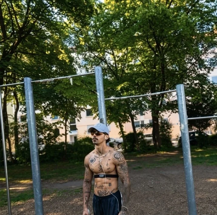 Brustloser Mann mit Tattoos, Sonnenbrille und Mütze, der im Park steht, während er Musik hört. Im Hintergrund sind grüne Bäume und eine Mehrfamilienhaus-Front.