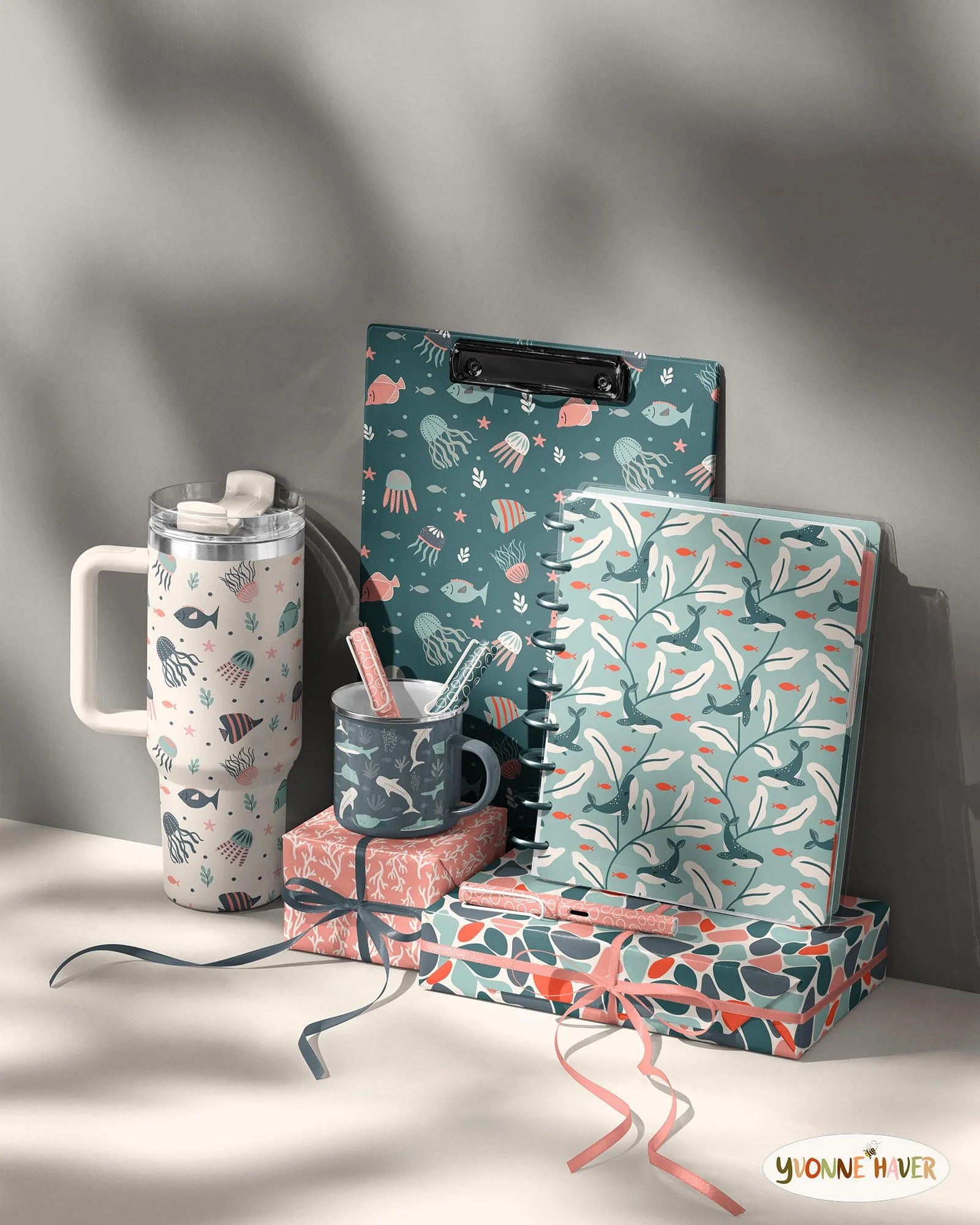 A collection of ocean-themed stationery and gift items, including a tall mug, a spiral-bound notebook, and wrapped presents, all decorated with fish, jellyfish, whales, and coral patterns in shades of blue, pink, and white, with curled ribbons and a small logo reading 'Yvonne Haver' at the bottom right.