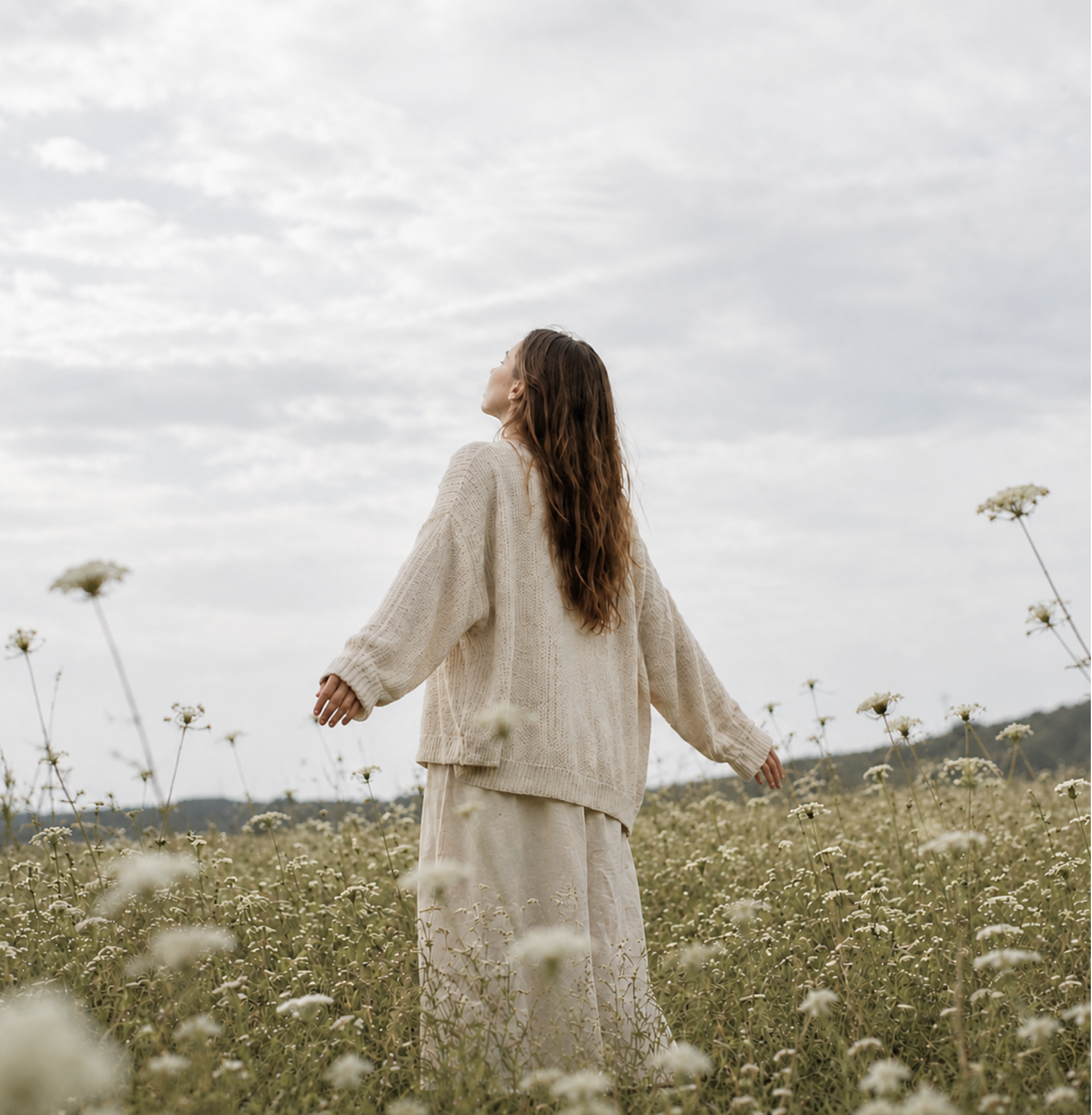 A woman with long wavy hair standing in a field of white flowers, facing upward with her eyes closed, wearing a cream-colored sweater and long skirt under cloudy skies.