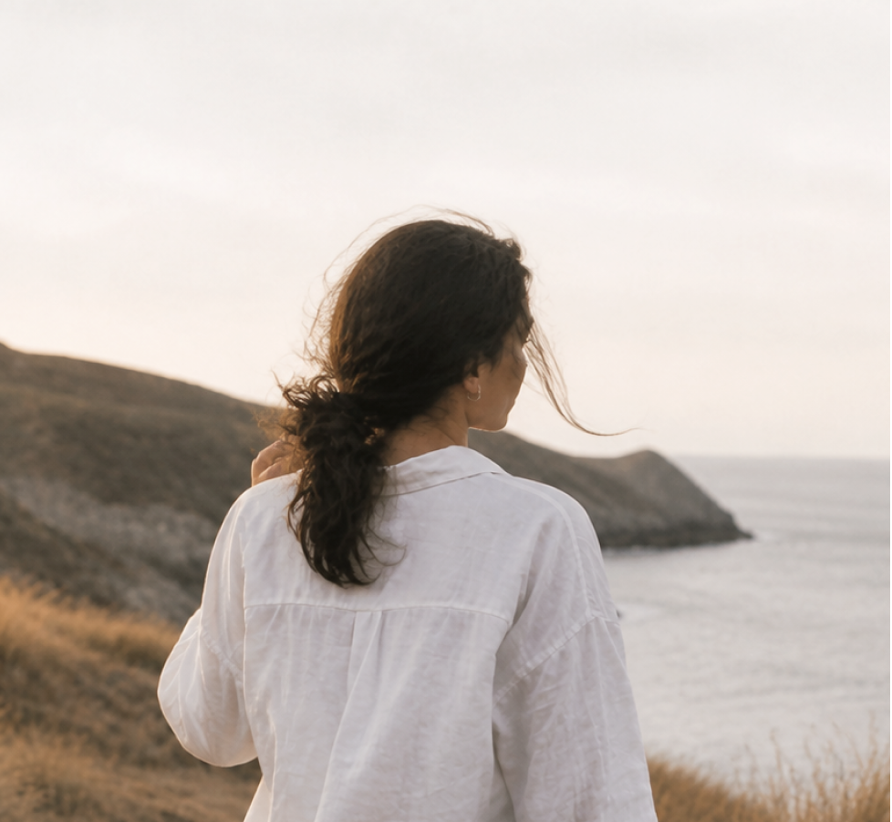 Woman with dark curly hair wearing a white shirt facing a coastal landscape at sunset with cliffs and water
