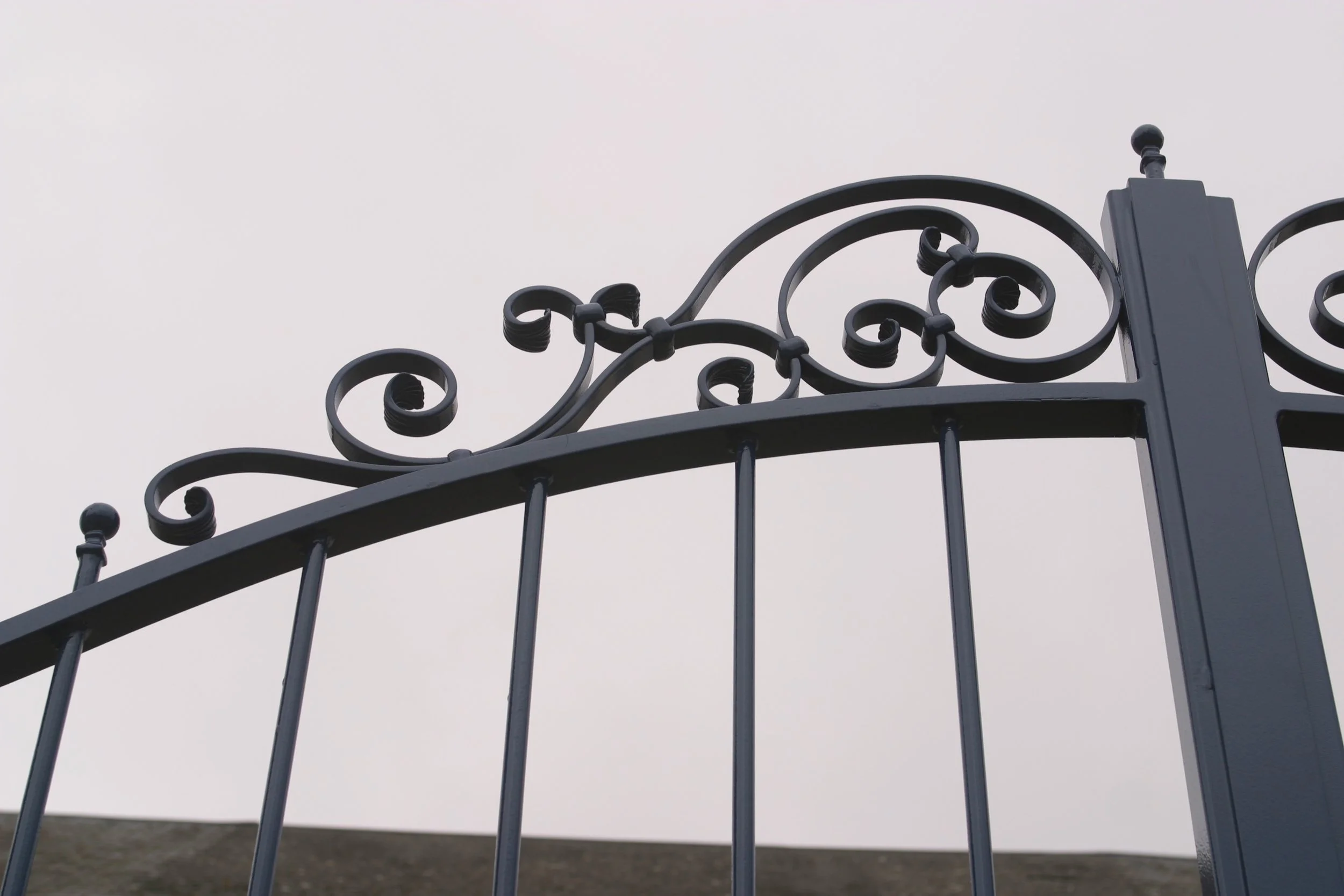 Une clôture en fer noir avec des décorations en spirale et boule de métal au sommet, sous un ciel gris