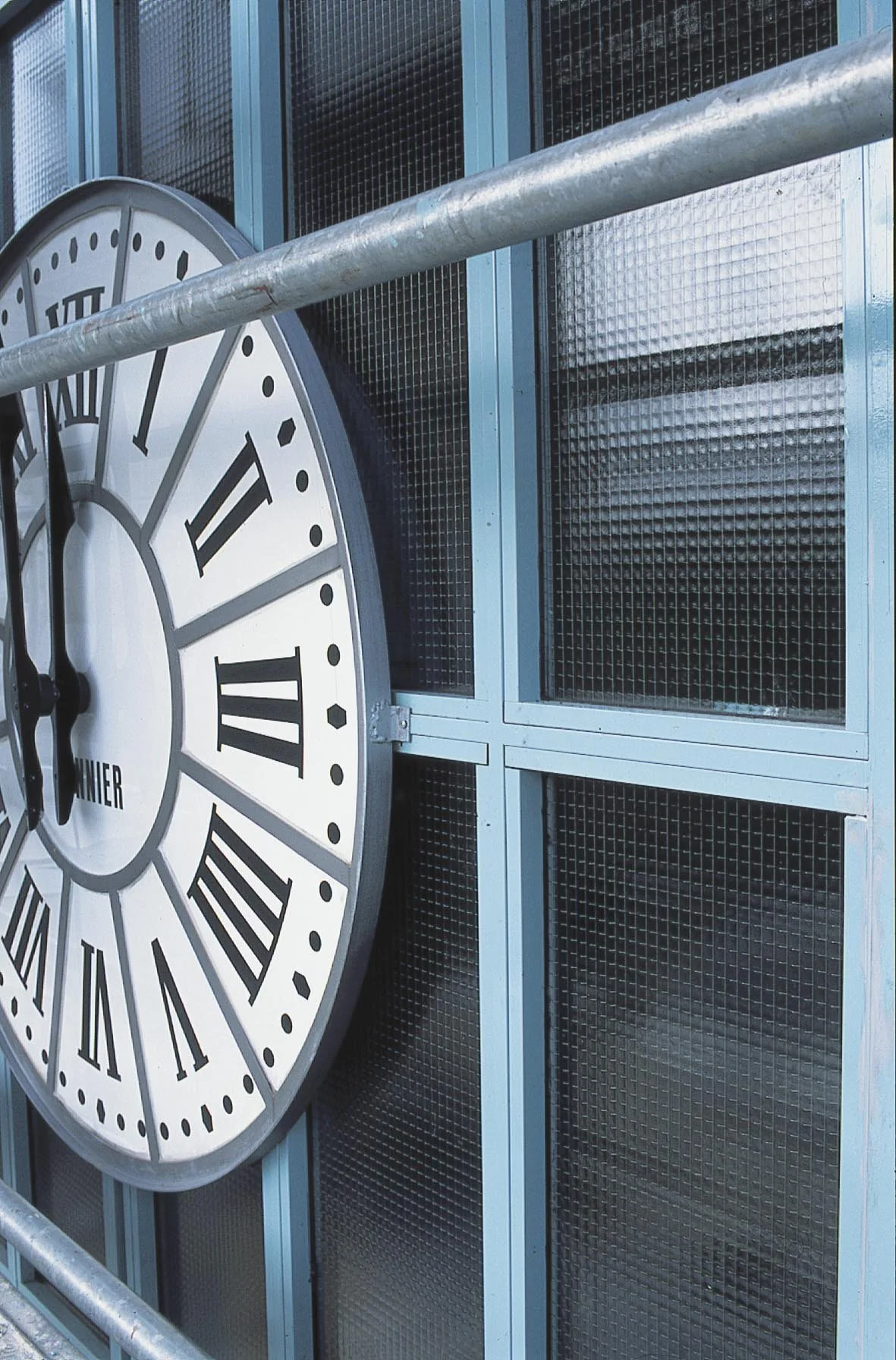 Horloge de grande taille avec chiffres romains, fixé sur une façade métallique entourée de grilles et de barres en acier.