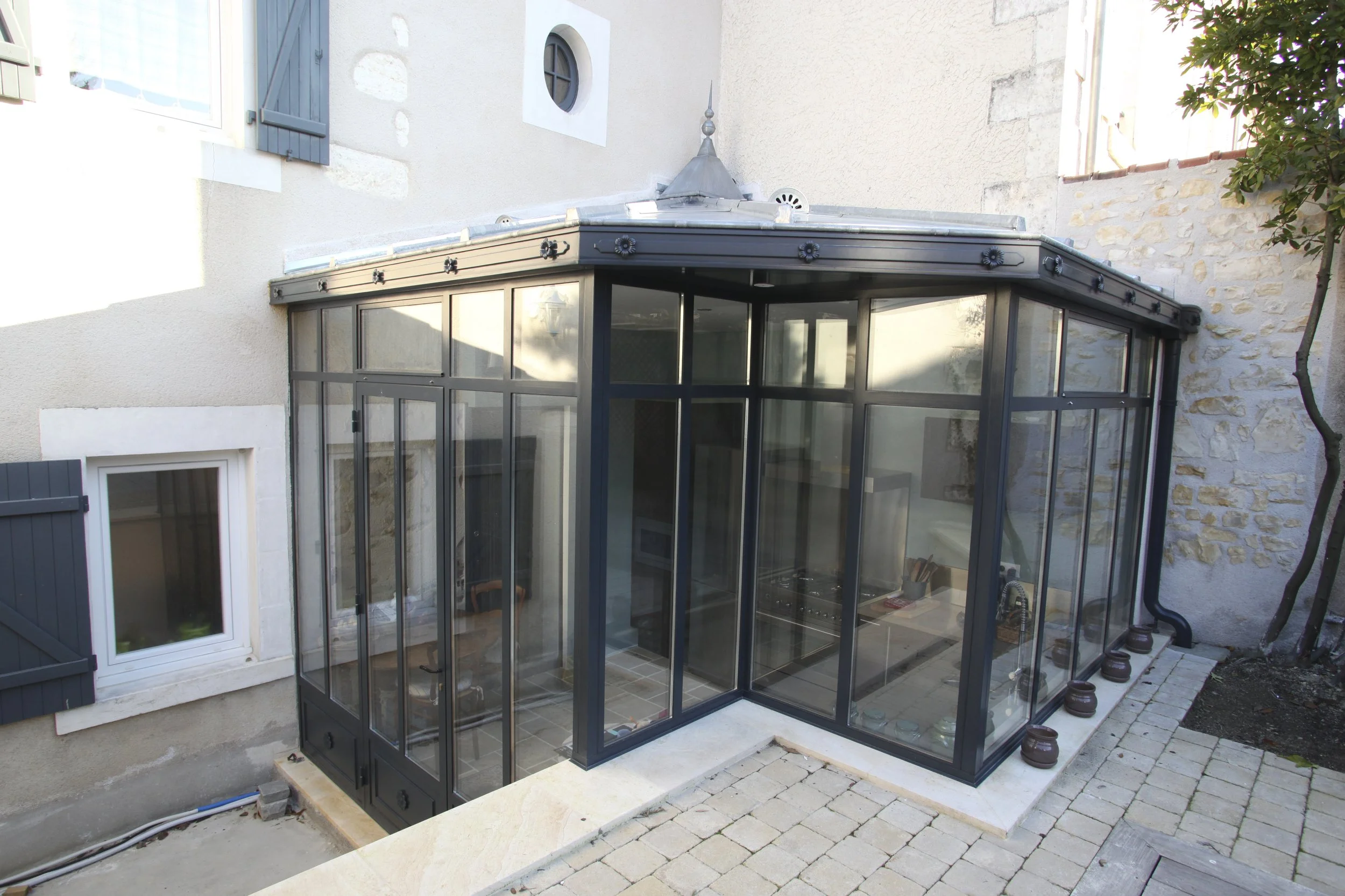 Une véranda en verre attachée à une maison en pierre, avec un sol en carrelage et plusieurs pots de fleurs alignés à l'extérieur.