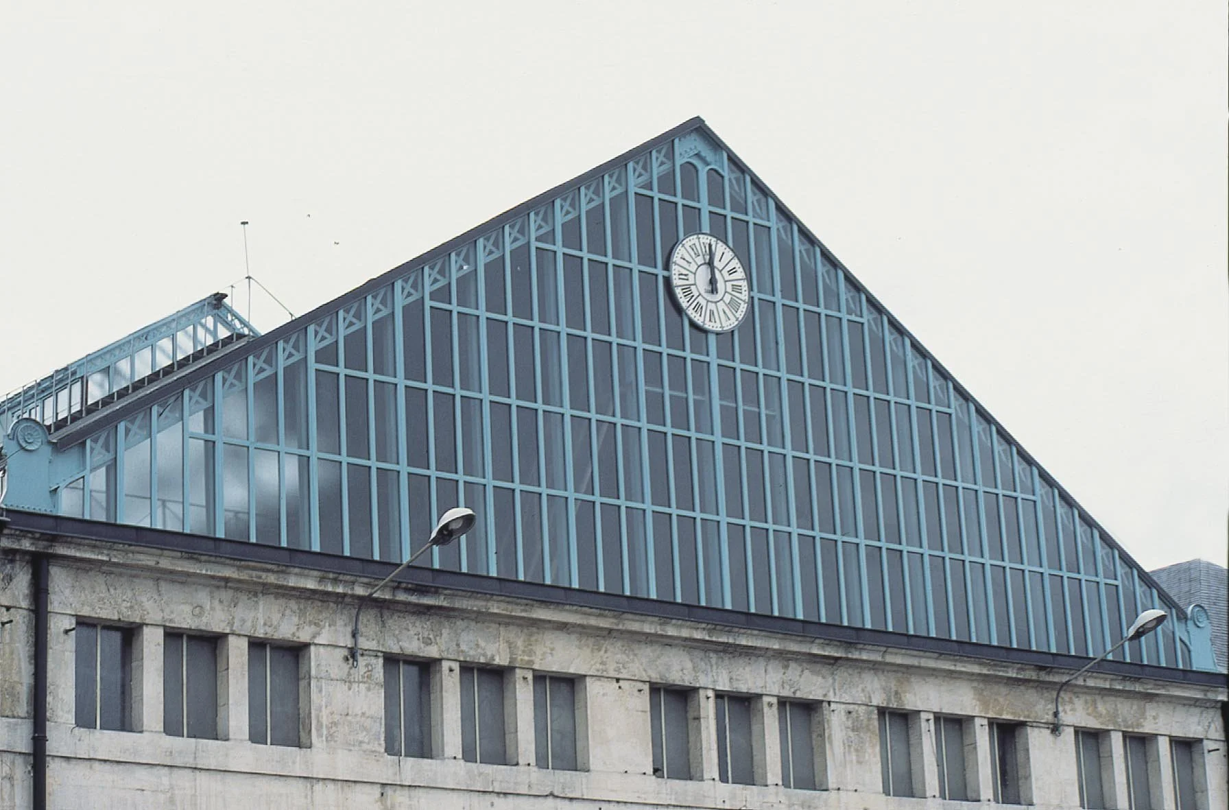 Un bâtiment moderne avec une façade en verre et une horloge murale. Le ciel est gris et il y a des lampadaires devant le bâtiment.