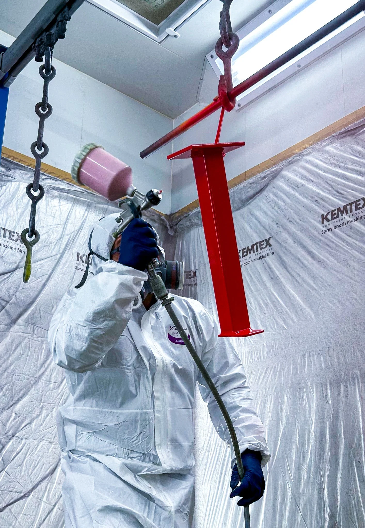 Une personne en combinaison de protection utilise un pistolet pulvérisateur pour appliquer une finition rouge sur une pièce en métal suspendue. La pièce est fixée à un support métallique vertical dans une cabine de peinture