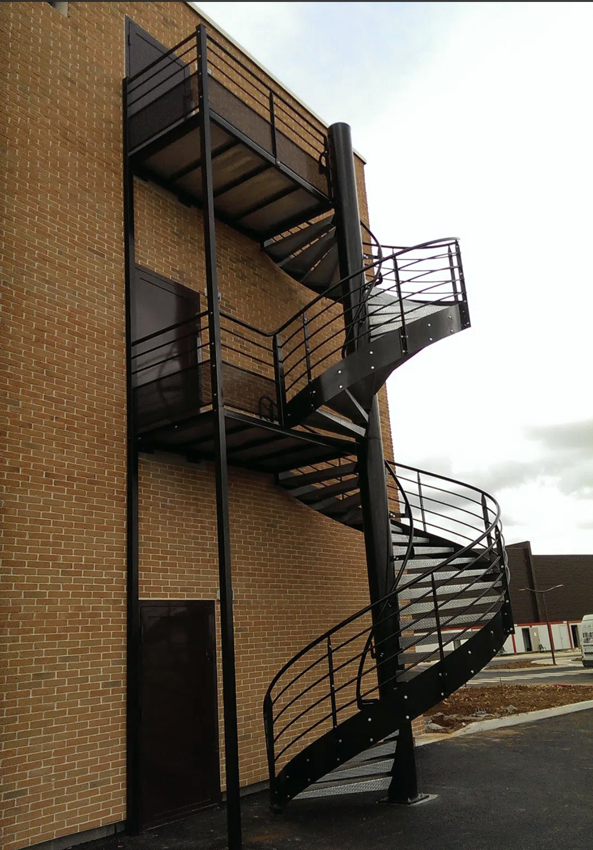 Escalier extérieur métallique en spirale attaché à un mur en briques, avec une plateforme au sommet, dehors par temps nuageux.