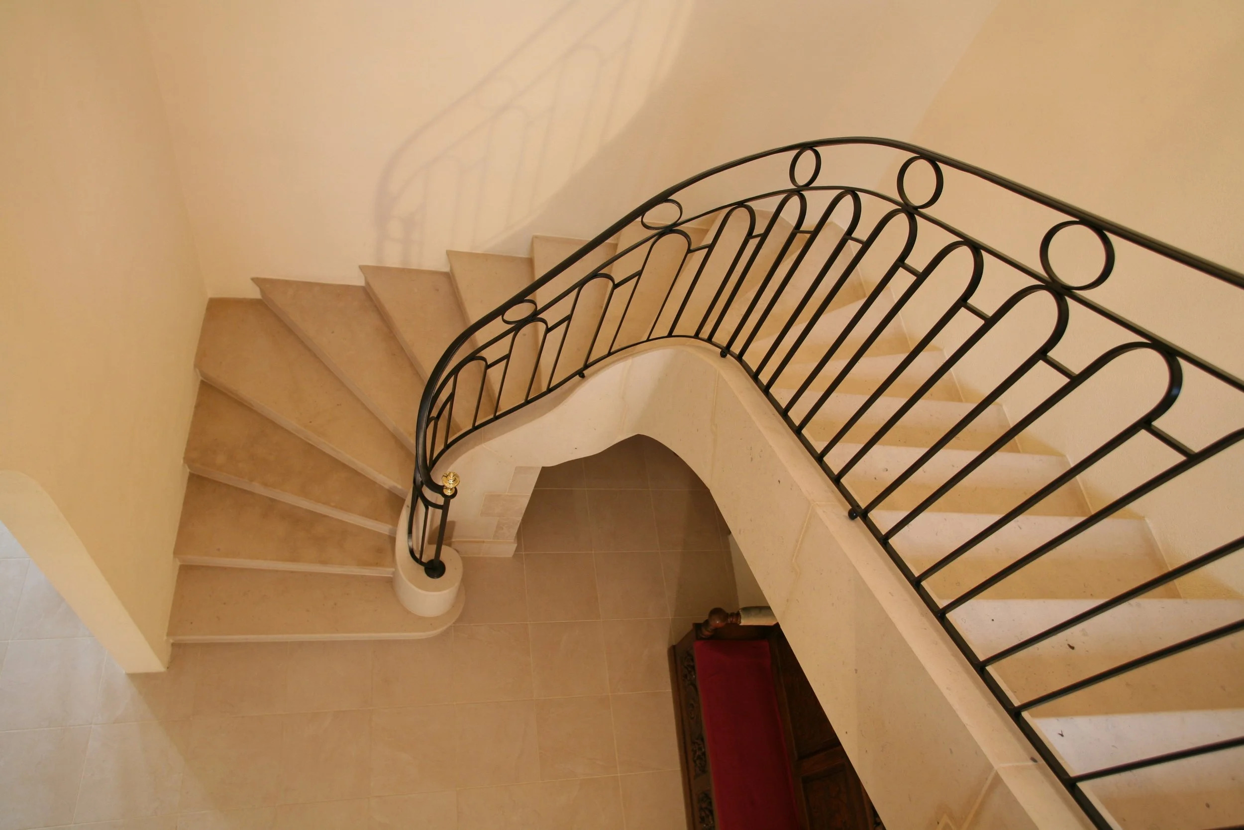Vue d'une cage d'escalier avec des marches en marbre beige, un mur jaune clair, une rampe en métal noir avec cercles, et un tapis rouge à l'entrée du palier.