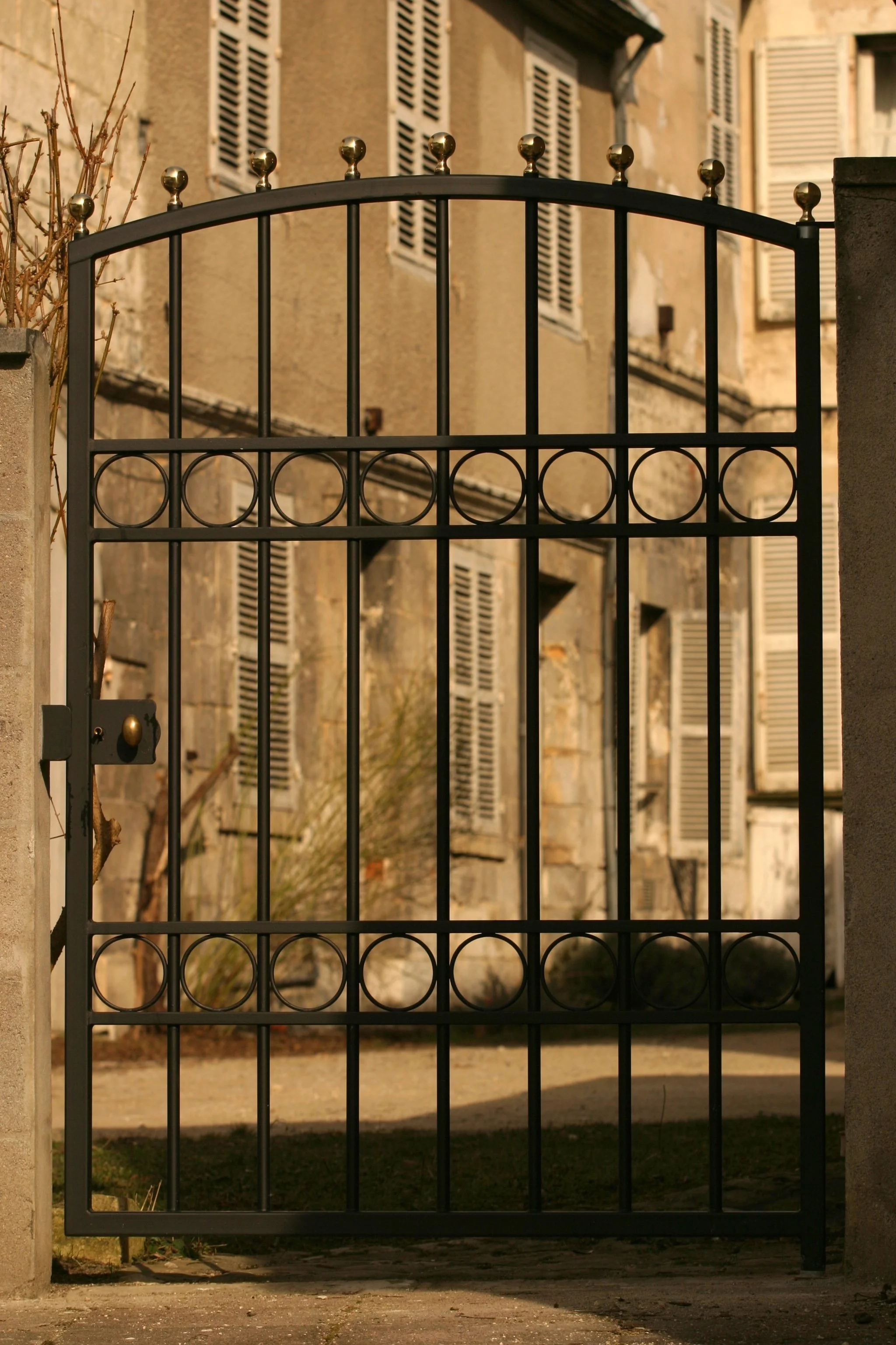Une porte de clôture en métal noir avec des décorations de boules en or à la sommet, en face d'un bâtiment ancien avec des fenêtres à volets en bois beige.