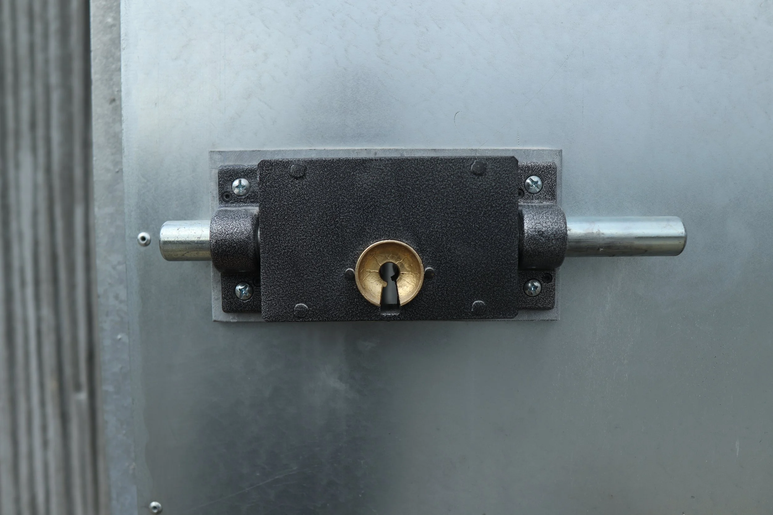 Caché derrière une surface métallique, un mécanisme de serrure avec une clé passe-partout. La serrure est fixée sur une plaque noire avec quatre vis, et un cylindre en métal dépasse du côté droit.
