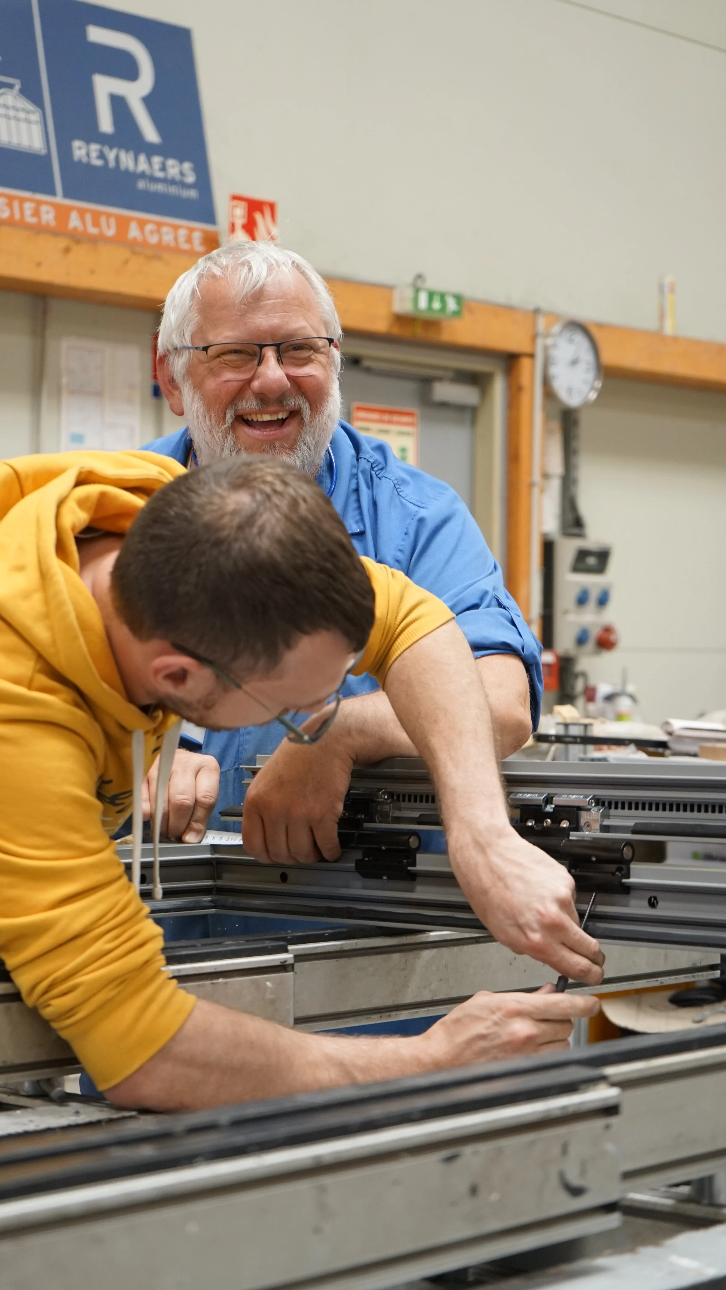 Deux hommes travaillent ensemble sur une machine en intérieur, l'un portant un pull jaune, l'autre une chemise bleue, tous deux souriants.