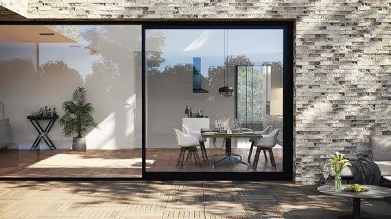 Salle à manger vue à travers une grande fenêtre avec des chaises blanches et une table, décor moderne avec mur en pierre et plantes