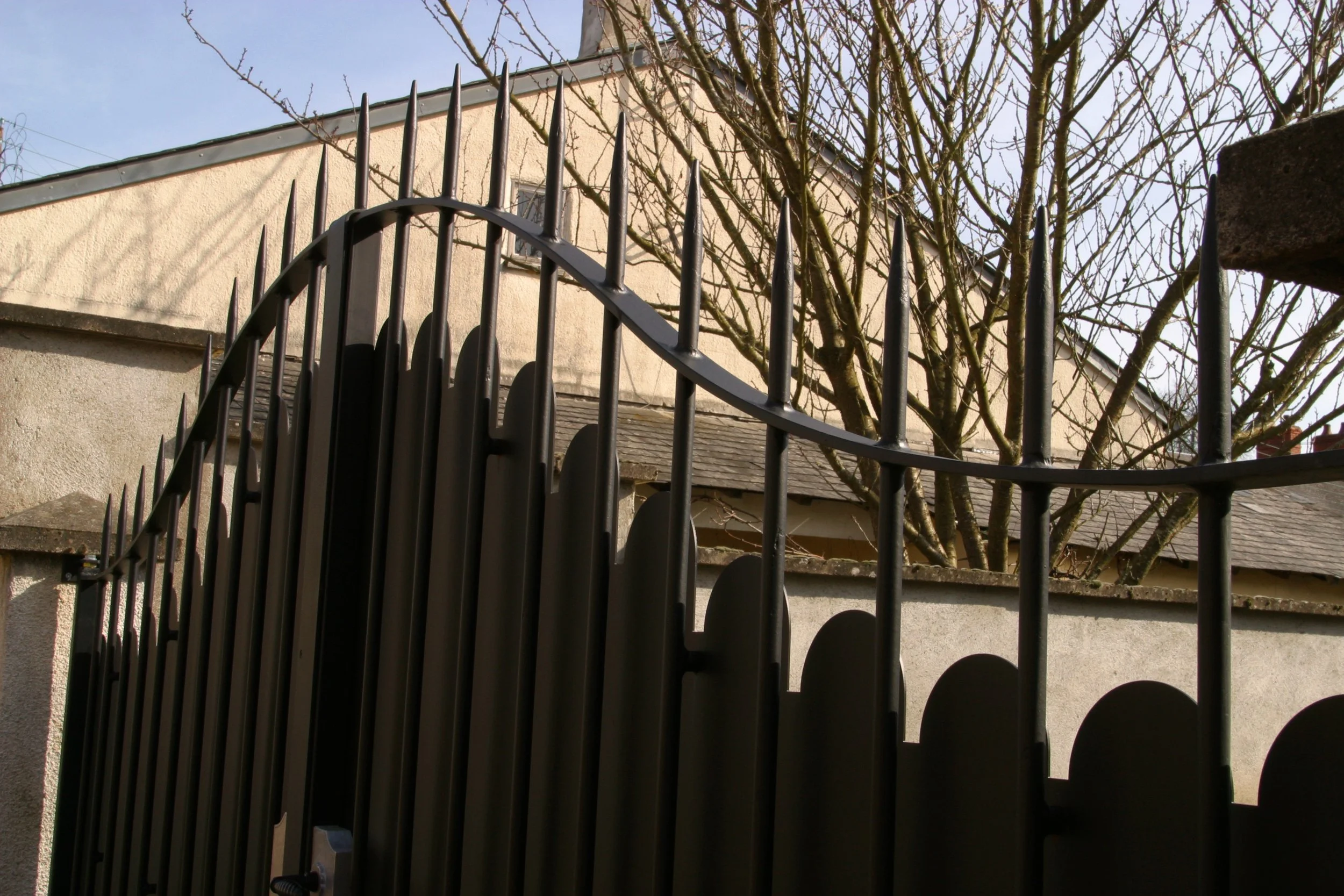 Une clôture en métal noire devant une maison beige avec un arbre sans feuilles en arrière-plan et un ciel bleu.
