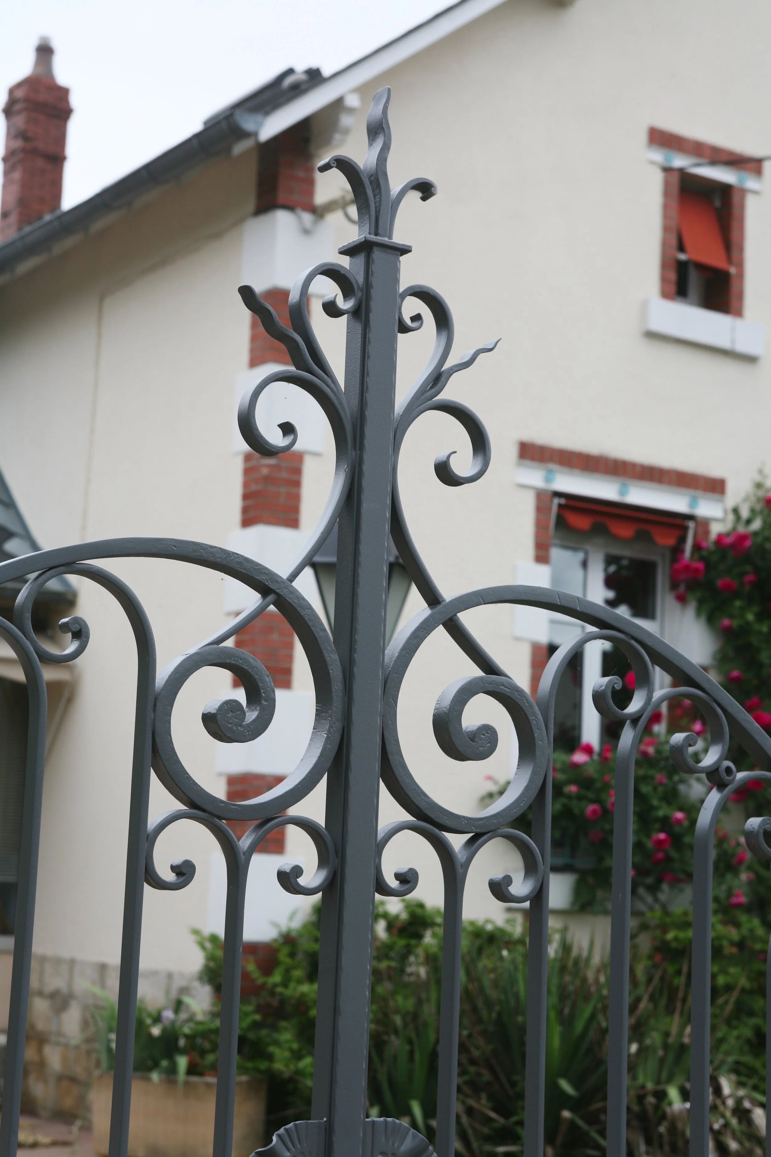Une clôture en fer forgé ornée de motifs torsadés en premier plan, avec une maison en arrière-plan, comprenant des murs beige, des fenêtres avec volets rouges, et un jardin fleuri.