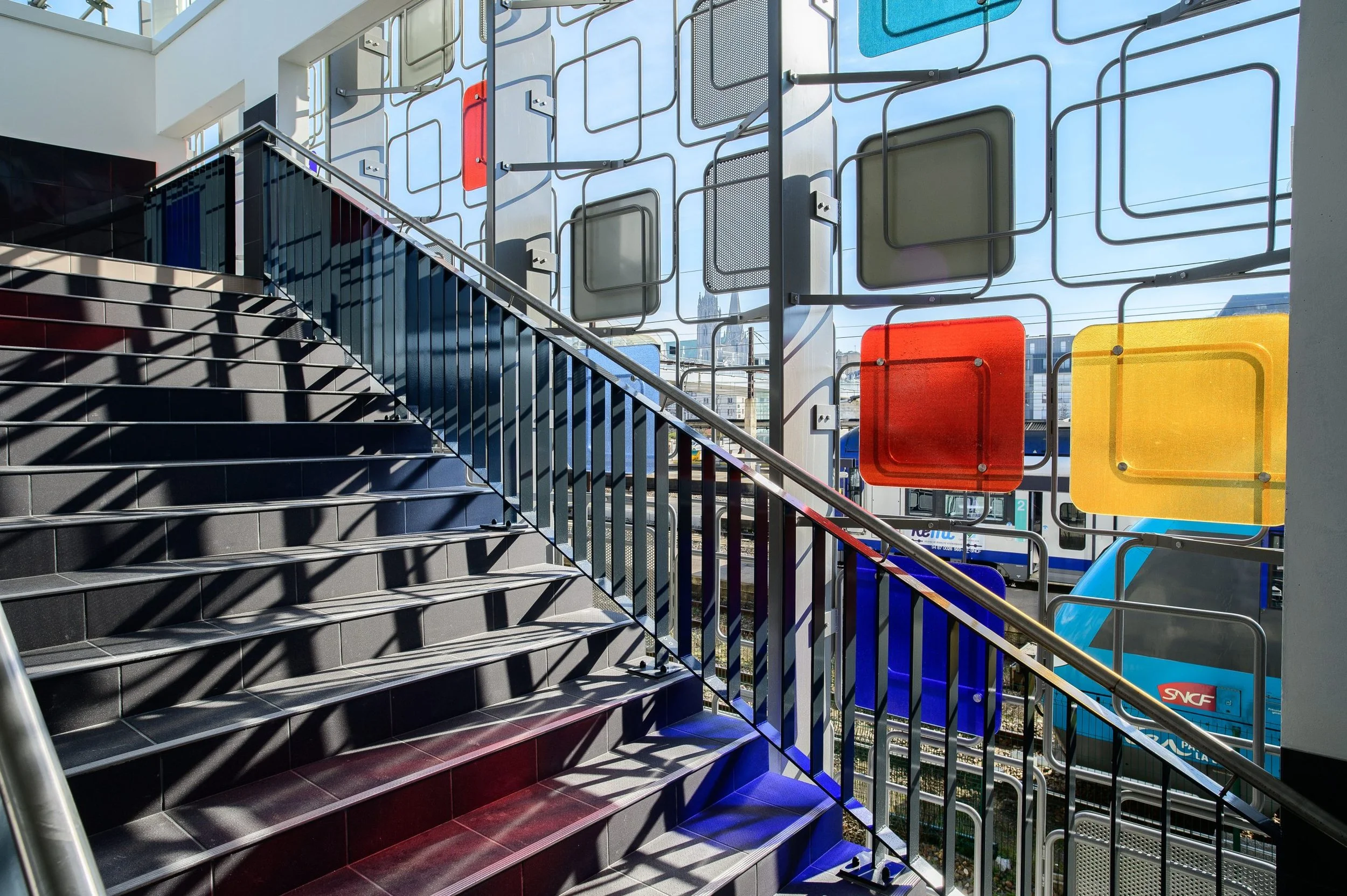 Escalier en métal noir menant à un espace intérieur lumineux, avec une façade en verre décorée par des formes géométriques colorées et une vue sur une station de métro ou train au travers du verre.