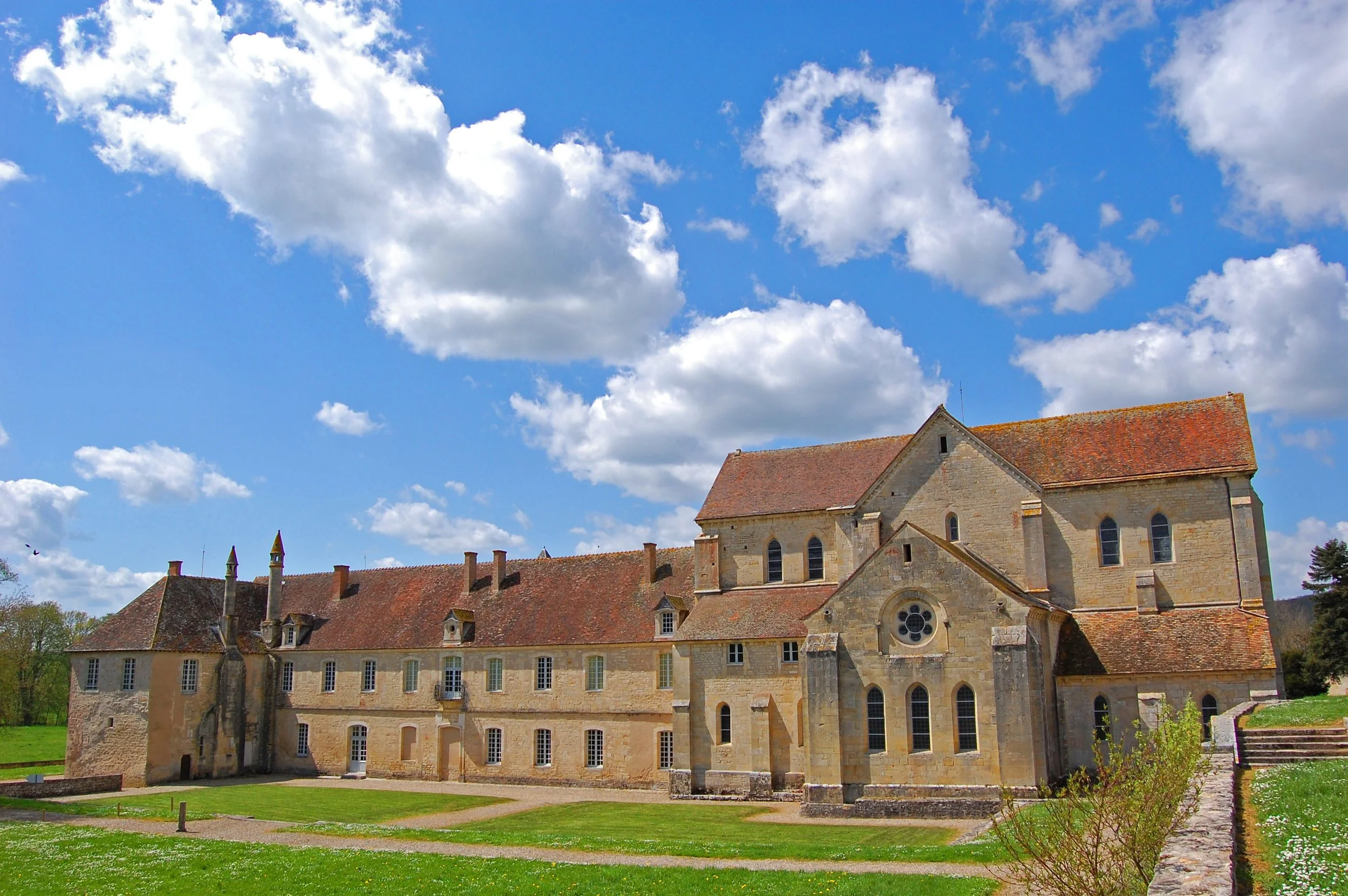 Vue d'un ancien bâtiment en pierre avec un toit en tuiles rouges, sous un ciel bleu avec des nuages blancs.