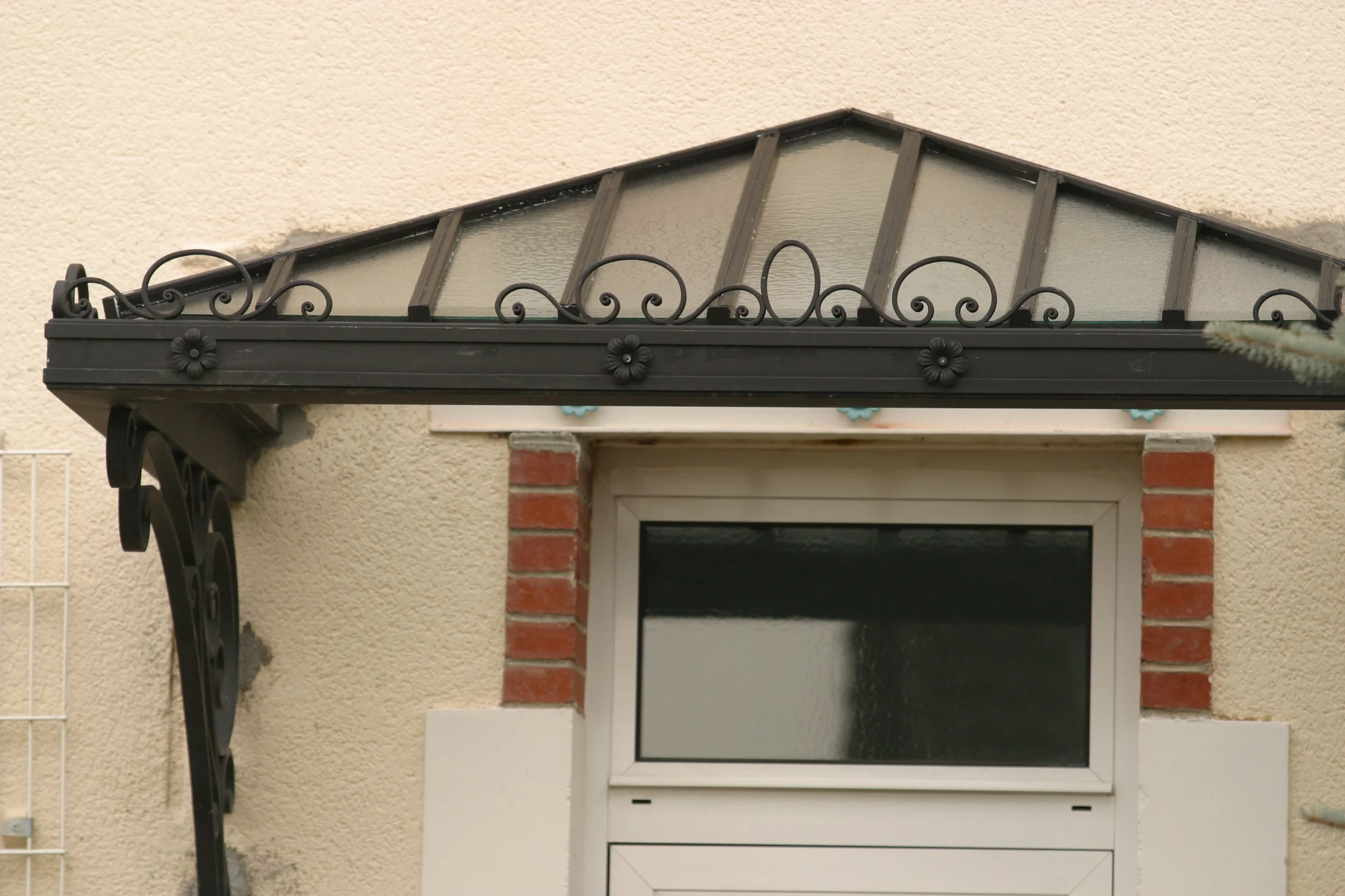 Fenêtre de toit avec cadre en métal noir et vitres en verre trempé, fixée au-dessus d'une porte d'entrée avec structure en brique rouge et encadrement blanc, sur un mur beige.
