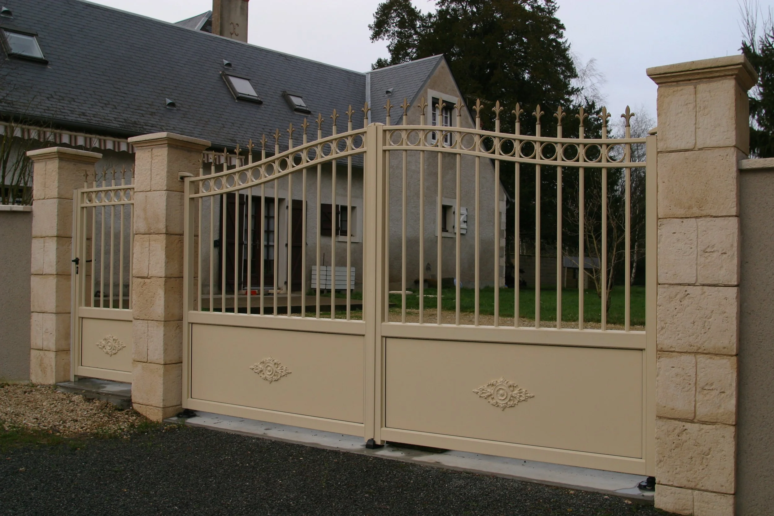 Une clôture en fer ornée de détails décoratifs, flanquée de piliers en pierre, en avant d'une maison avec un toit en ardoise et plusieurs fenêtres. Un arbre est visible en arrière-plan.
