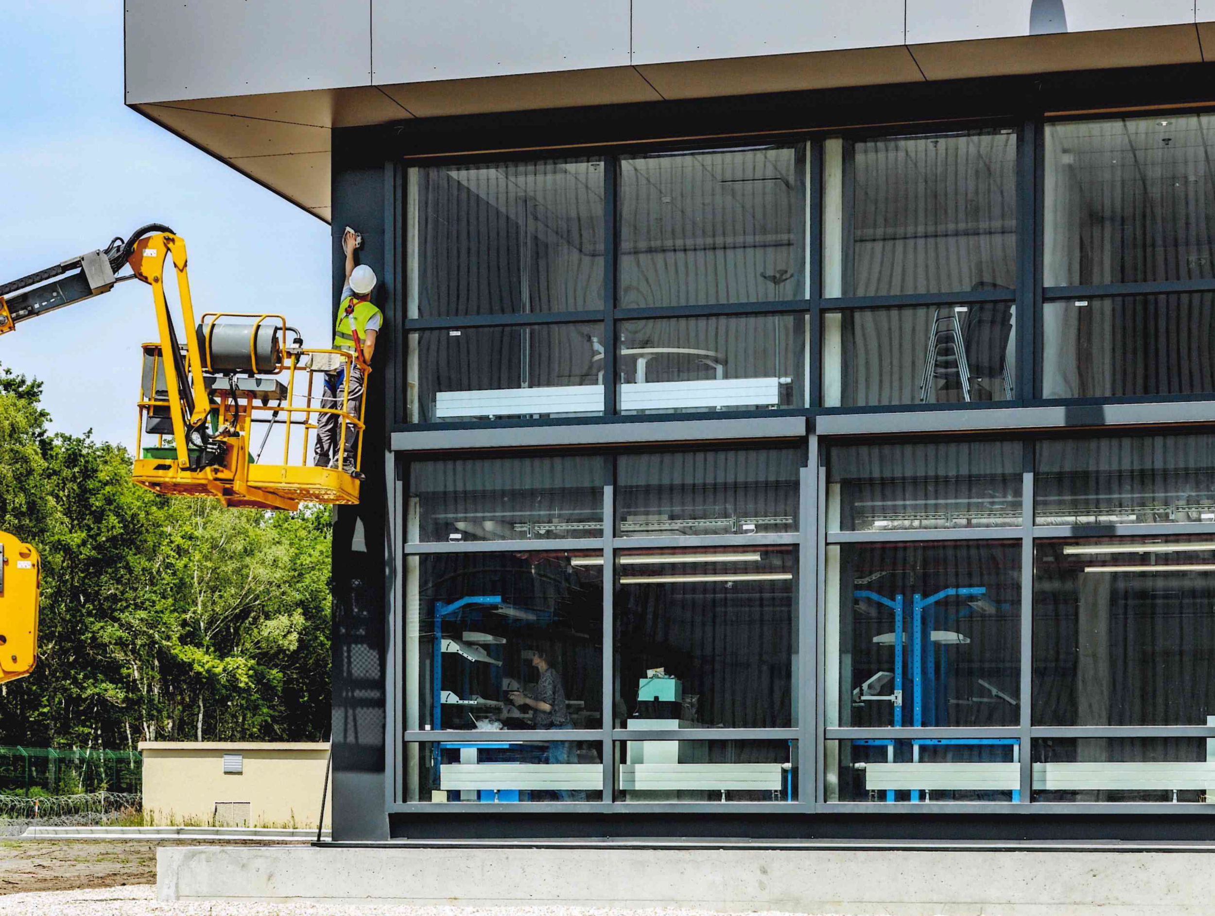 Un ouvrier en gilet de sécurité et casque travaille sur une façade d'un bâtiment moderne avec de grandes fenêtres en verre, utilisant une plateforme élévatrice.