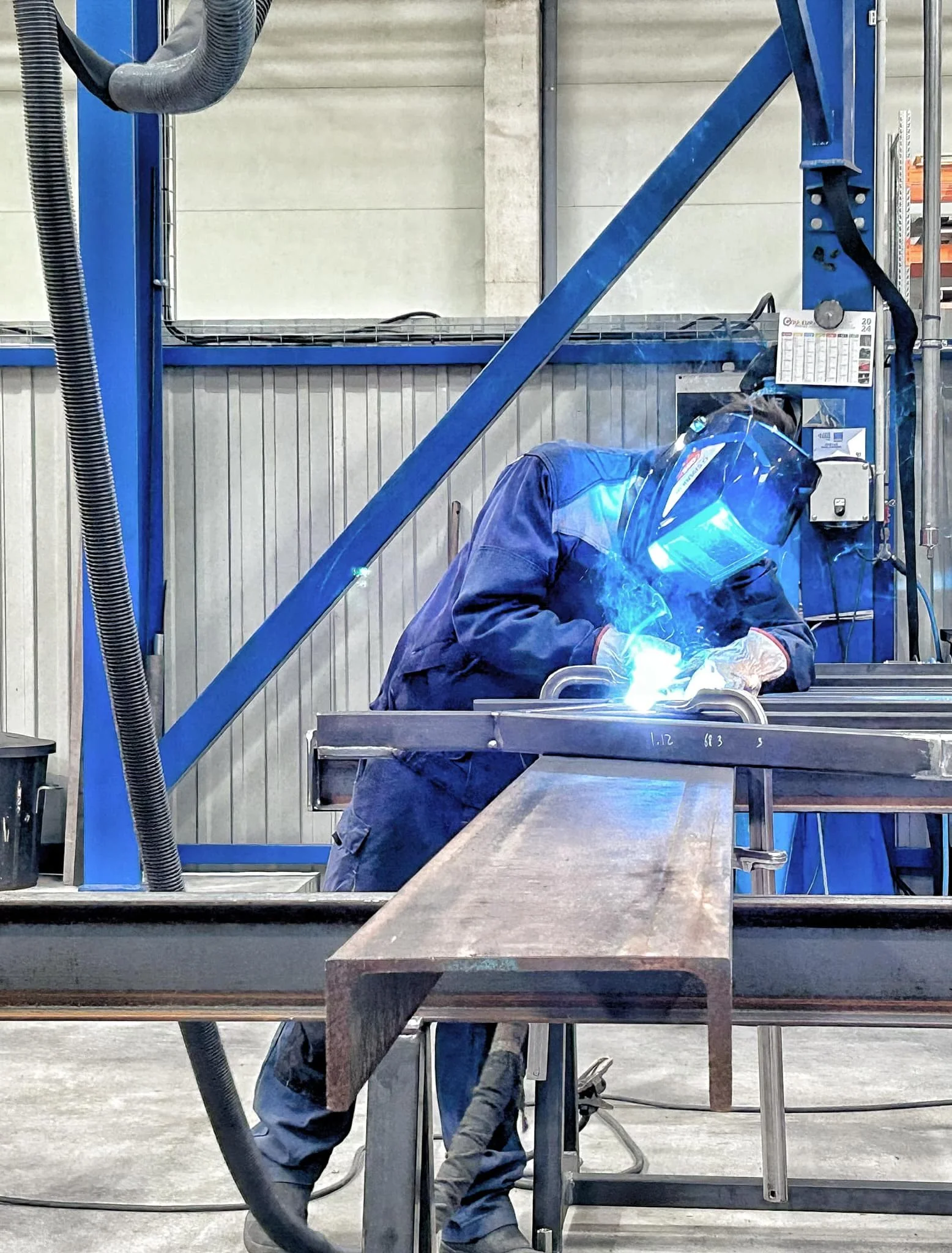 Un ouvrier en combinaison de travail et masque de soudage effectuant une soudure sur une pièce métallique dans une usine ou un atelier.