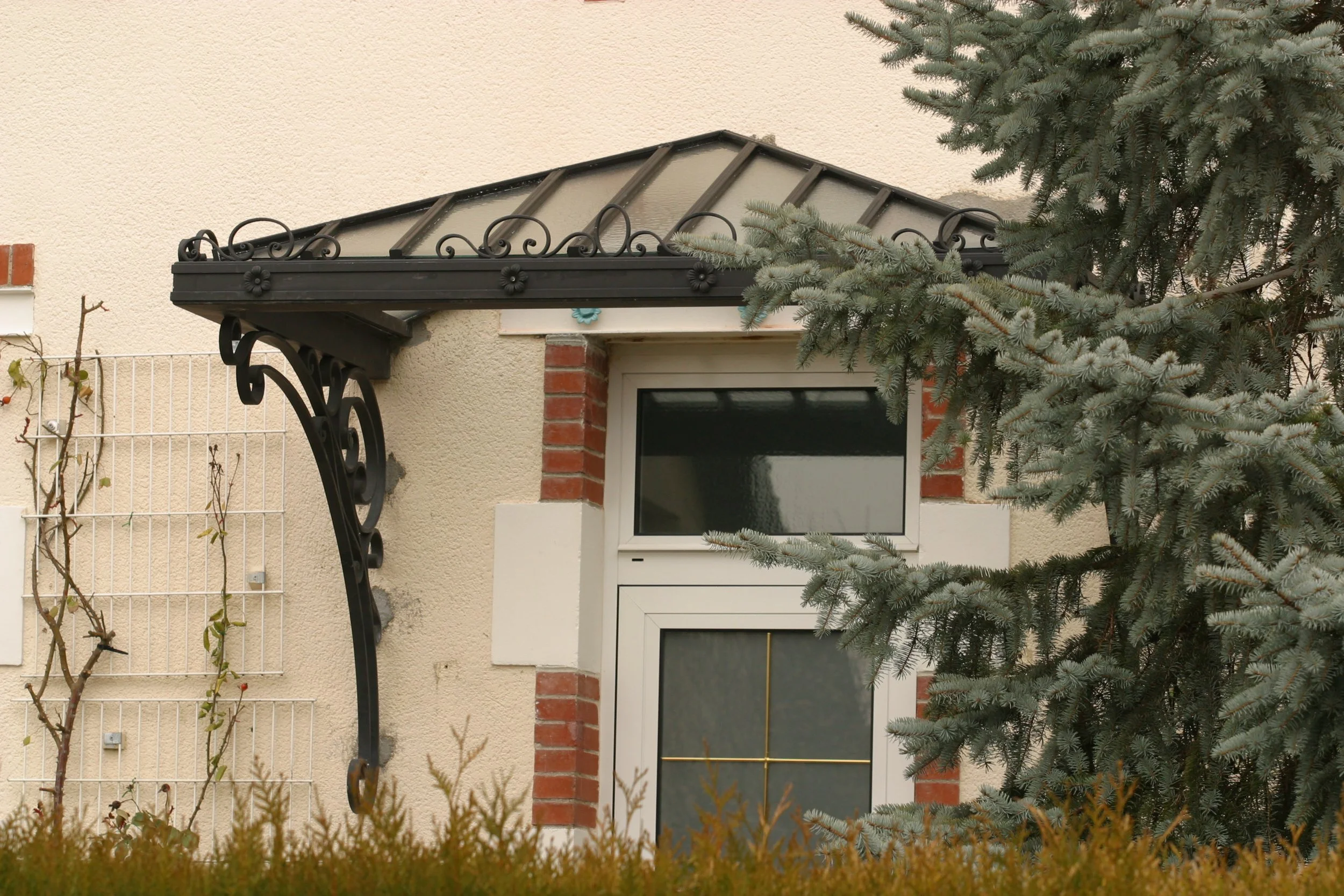 Vue partielle d'une maison avec porte-fenêtre, entourée de végétation et d'un arbre à aiguilles, avec un auvent en métal noir sur le devant.