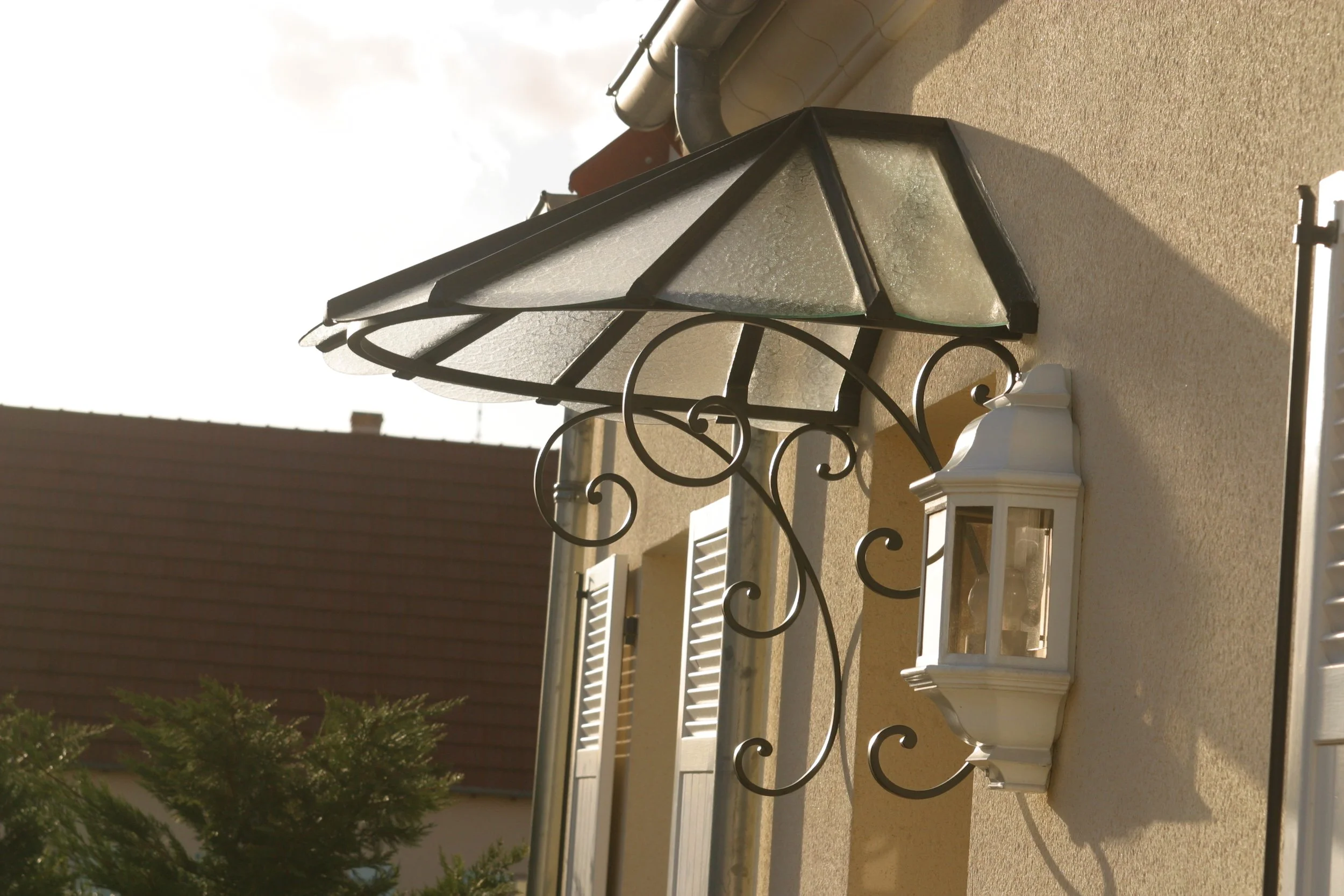 Une lanterne de balcon en pierre blanche avec une structure en métal noire, placée sur un rebord de balcon en pierre beige. En arrière-plan, il y a des fenêtres à volets blancs et un arbre vert, avec un ciel en partie couvert.