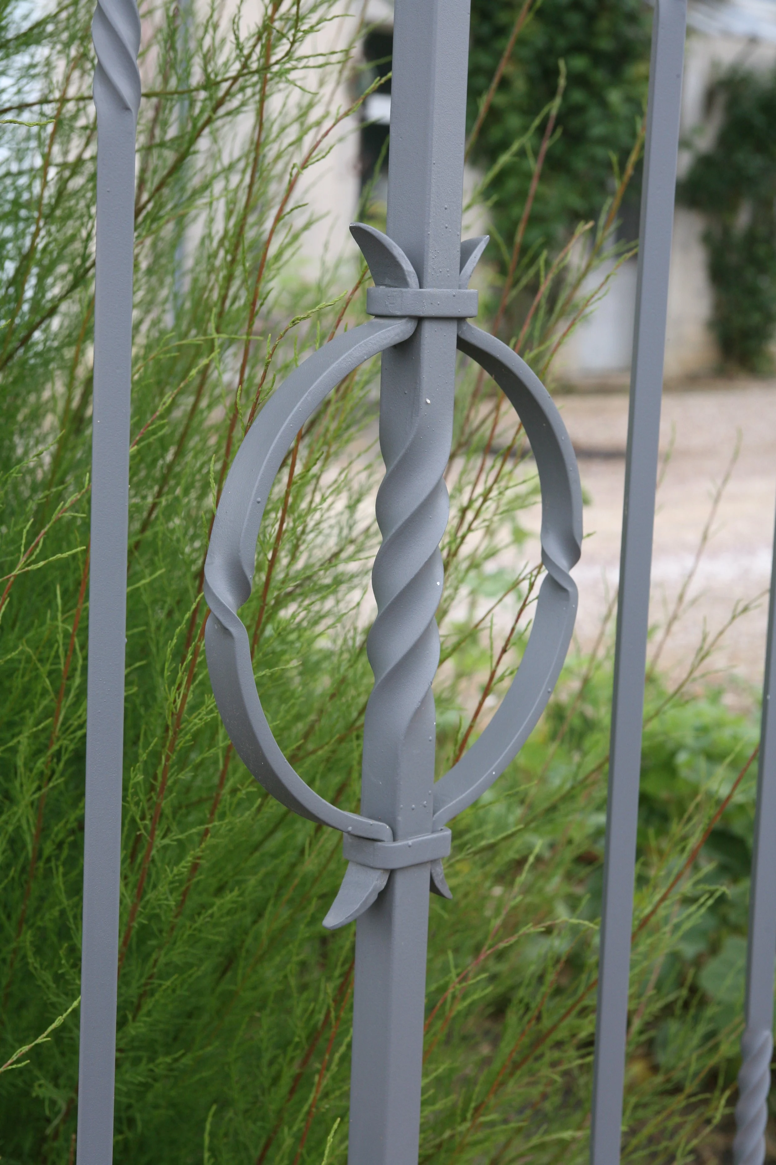 Une clôture en métal gris avec un motif en forme de cercle avec des branches incurvées, entourée de végétation verte