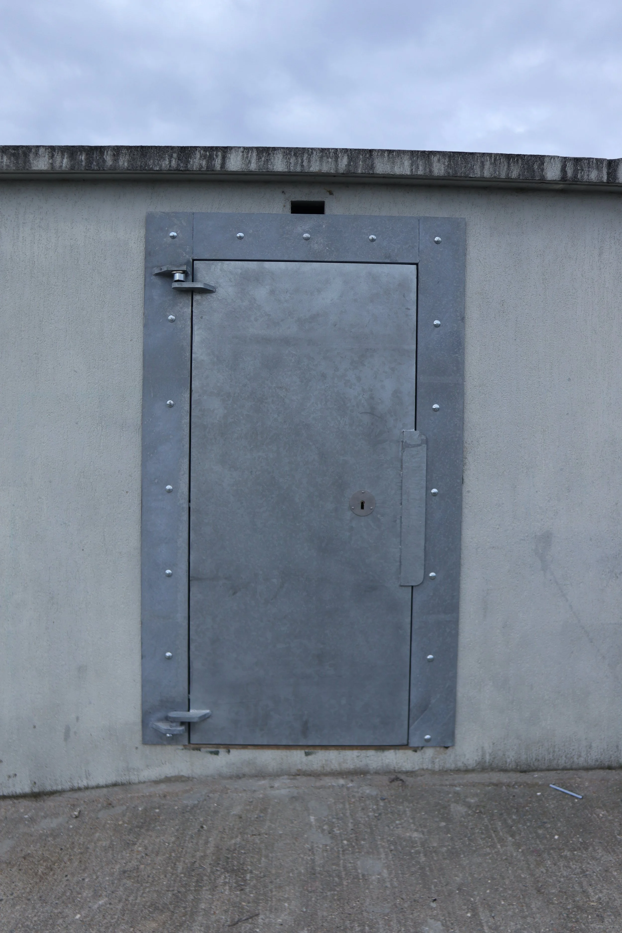 Une porte métallique grise montée sur un mur beige, avec un verrou et des rivets visibles, sous un ciel nuageux.