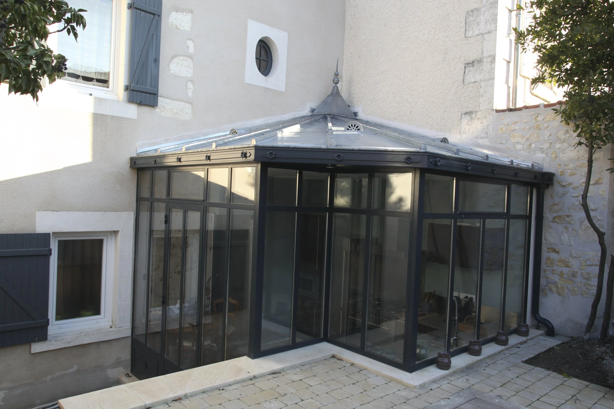 Une extension en verre noire de style conservateur avec des fenêtres rectangulaires et une toiture en métal, attachée à une maison ancienne en pierre avec des volets bleus et des murs en plâtre beige, située en extérieur avec quelques arbres et pavés