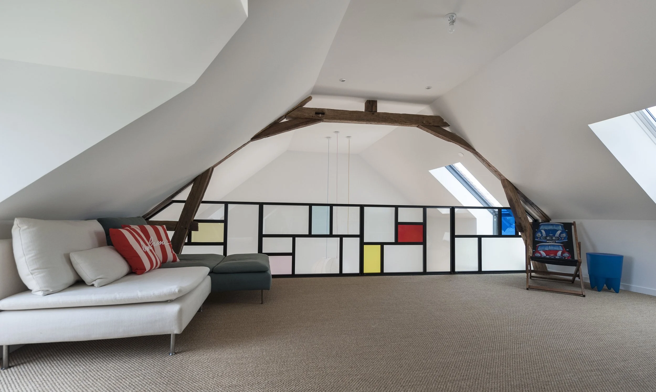 Salon mansardé avec canapé blanc, coussin rouge et gris, plancher en moquette, murs blancs, poutres en bois apparentes, fenêtres de toit, séparation en verre coloré et bois, coin enfants avec jouets et chaise en bois.