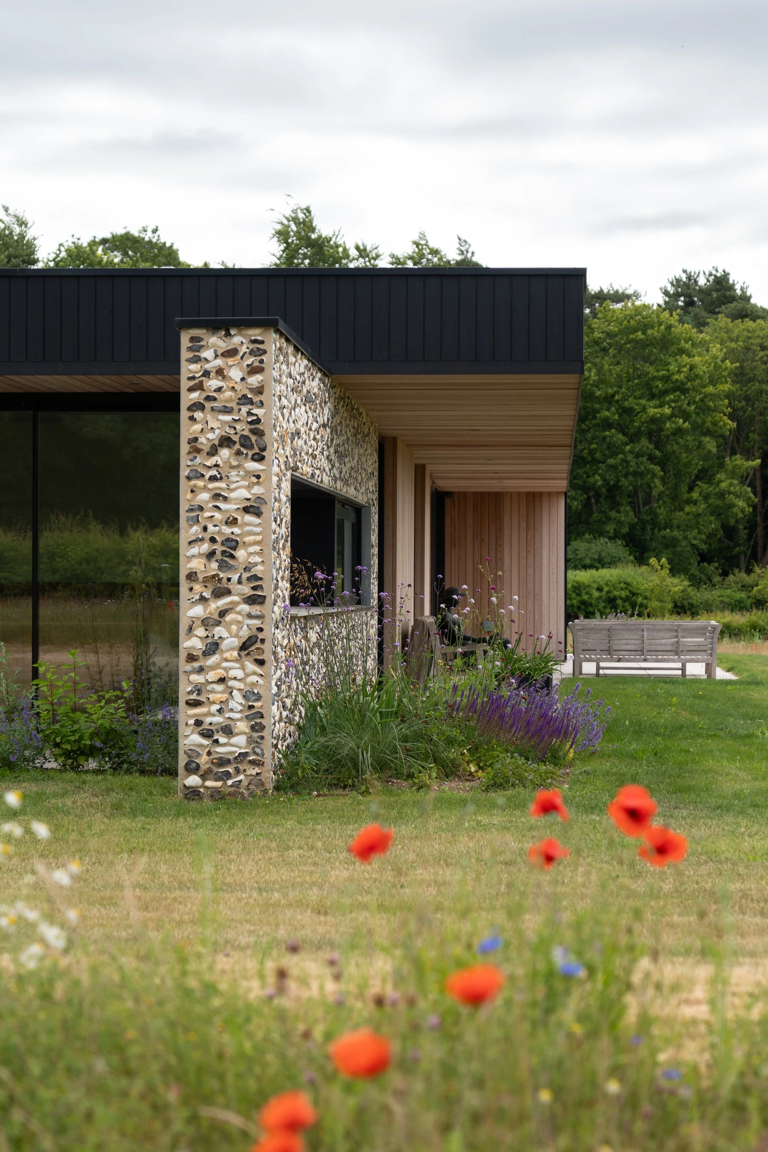 Modern house with stone and wooden exterior walls, surrounded by a garden with colorful flowers, and a wooden bench under the overhang.