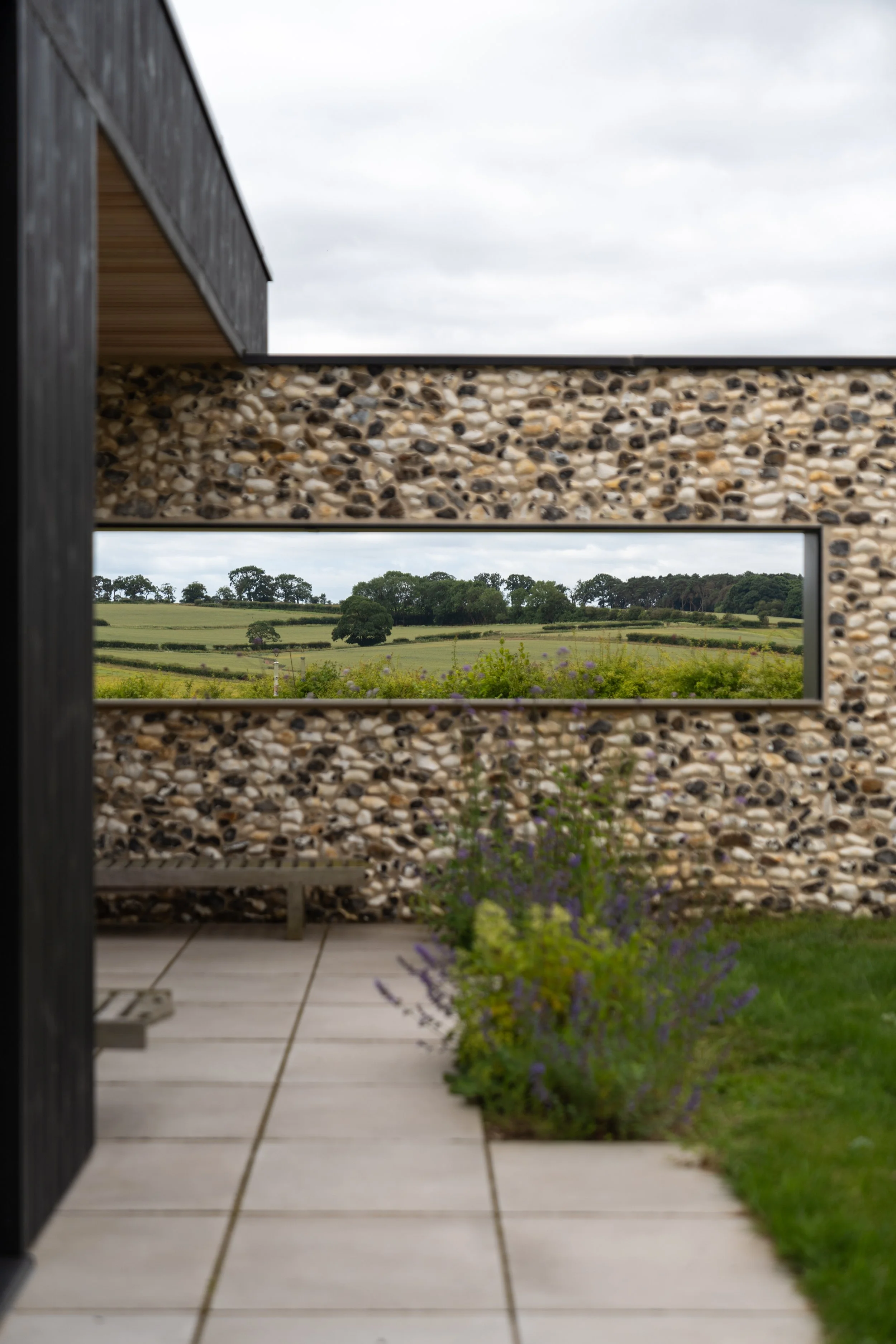 View of a garden through a rectangular opening in a stone wall, with an outdoor patio area and greenery in the background under a cloudy sky.