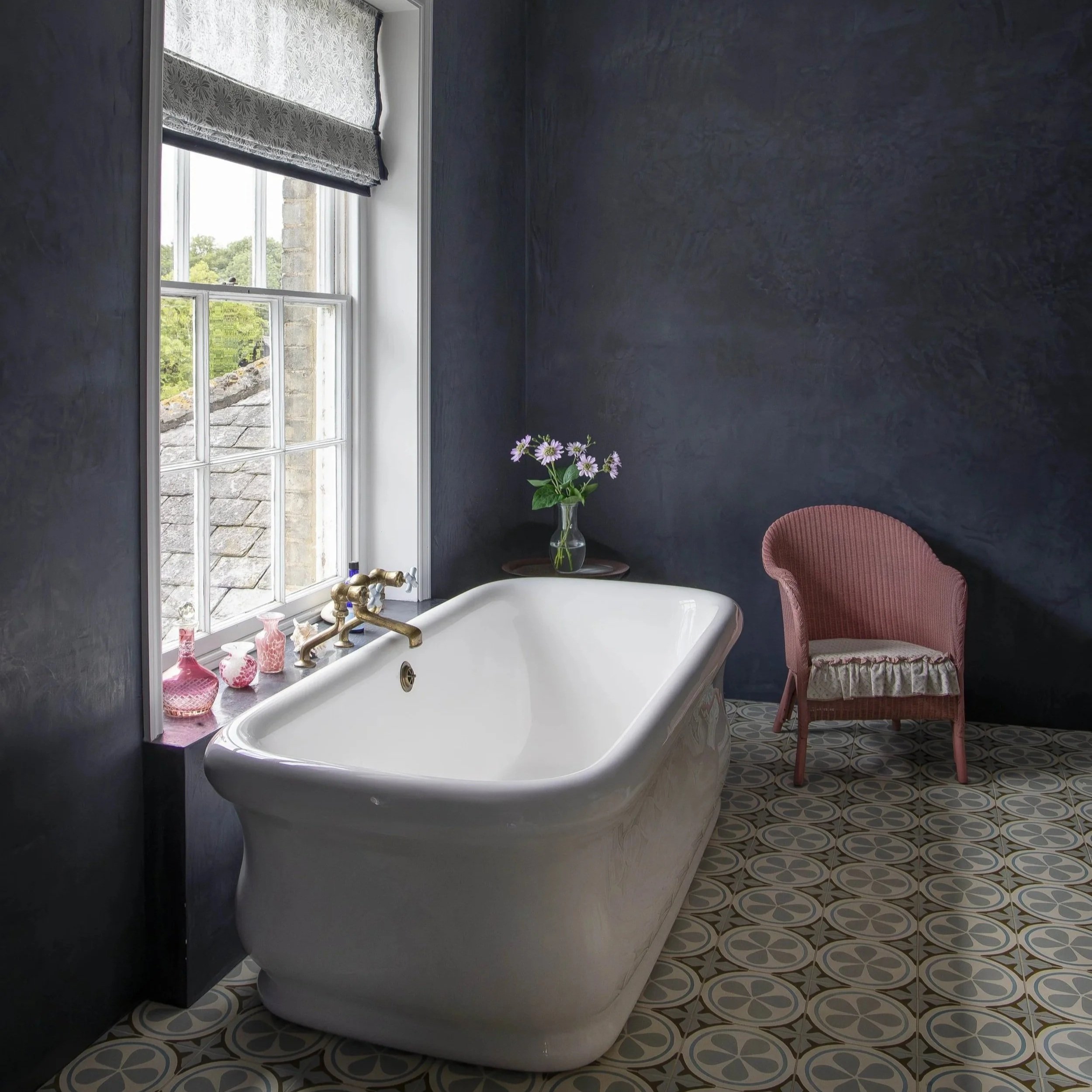 A bathroom with a vintage clawfoot bathtub near a window, decorated with pink vases and flowers, dark textured walls, and a patterned tiled floor, with a pink cushioned chair beside the tub.