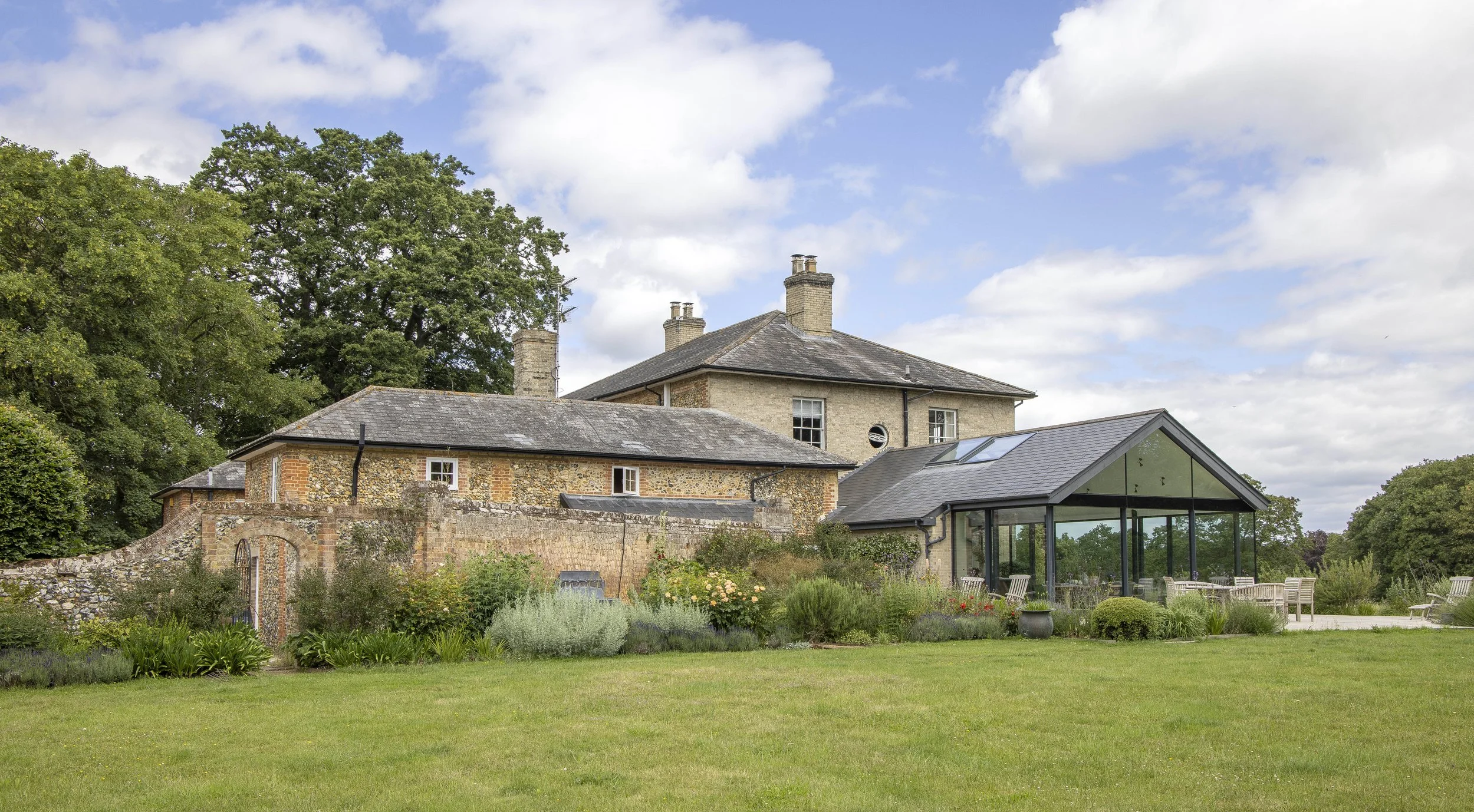 A large stone house with multiple chimneys, surrounded by trees and a garden, with a modern glass extension and outdoor seating area.